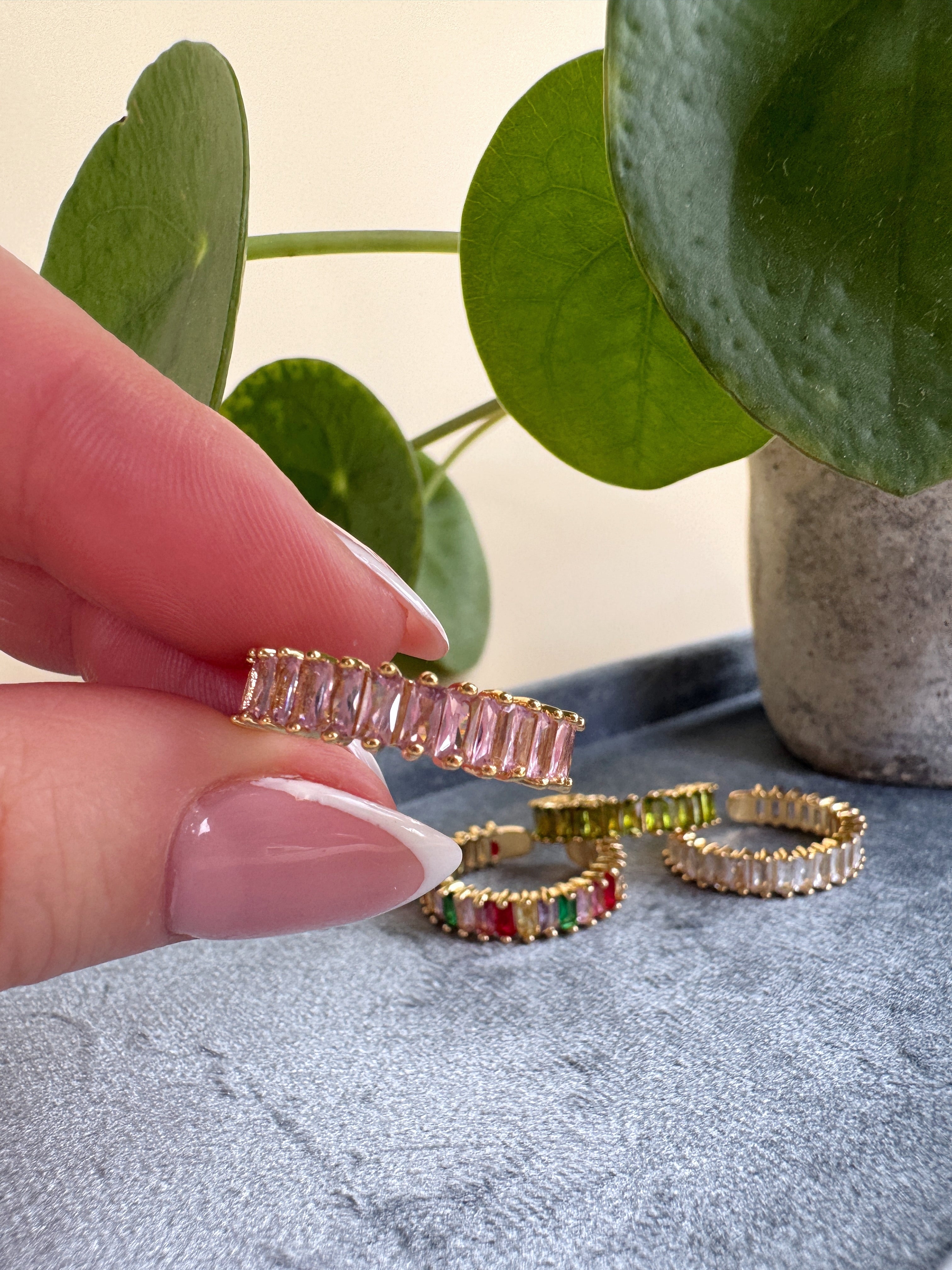 Gold rings with pink gemstones held by a hand against a neutral background with greenery.