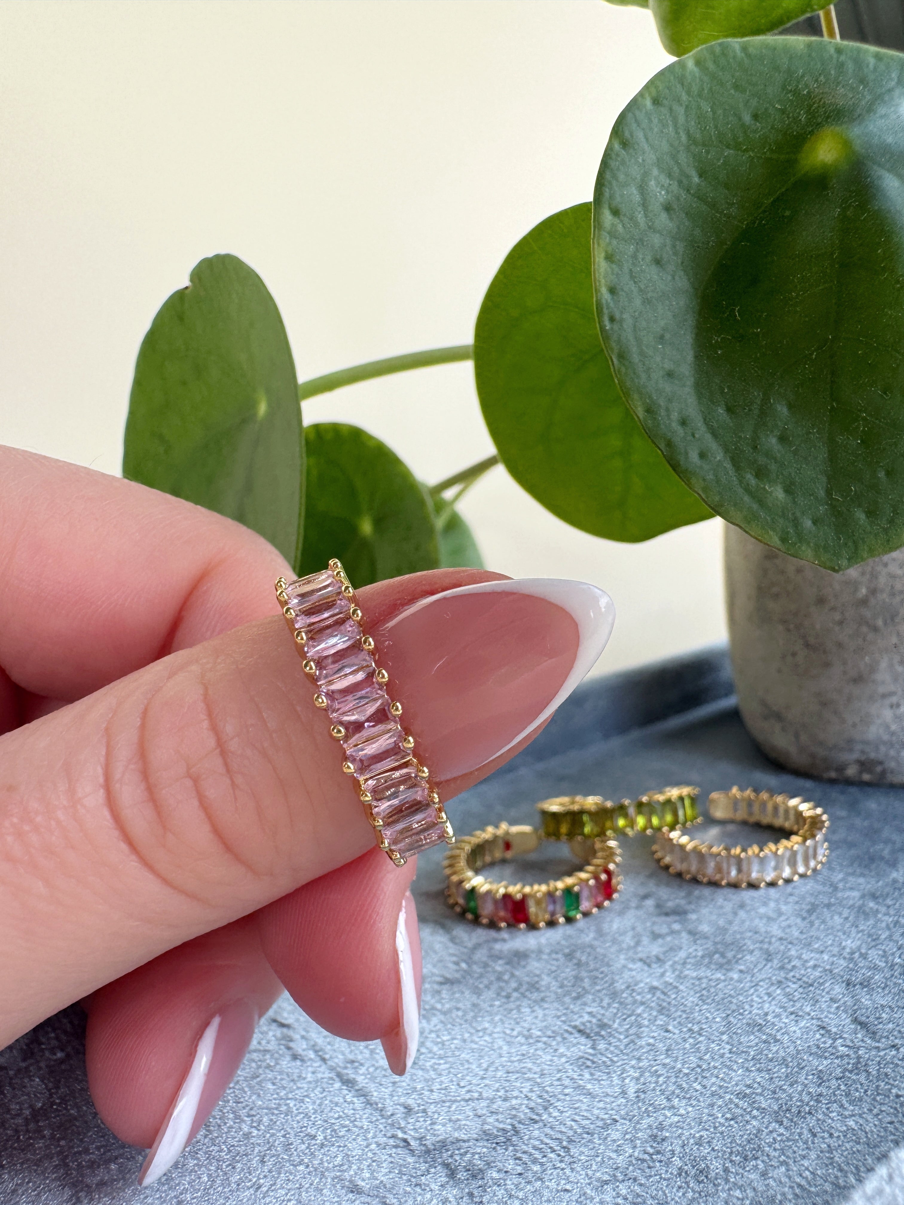 Hand holding a pink gemstone size adjustable ring with other rings on a surface and plant in the background