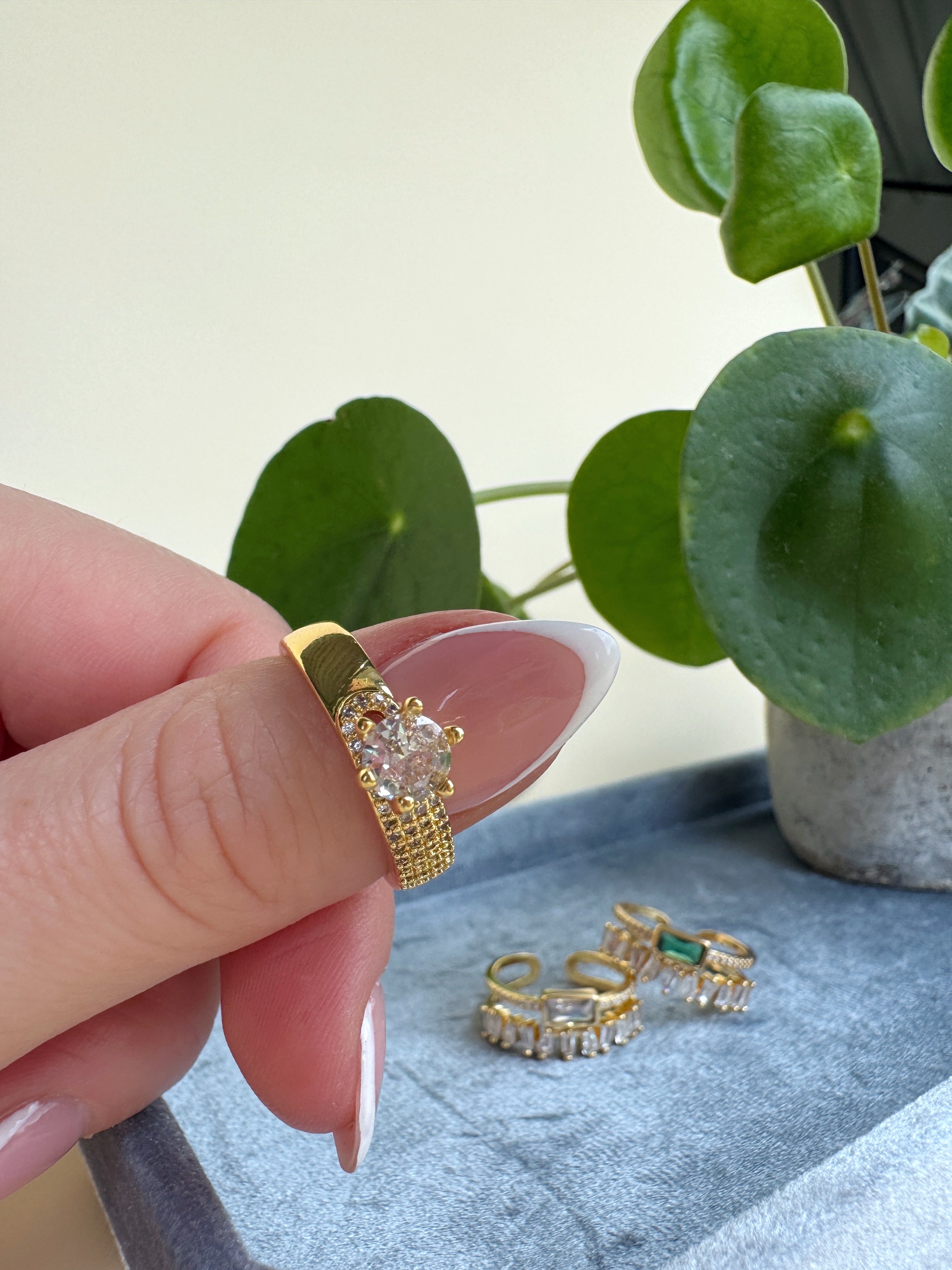 Gold ring with a diamond held by a hand, with more rings on a surface in the background.