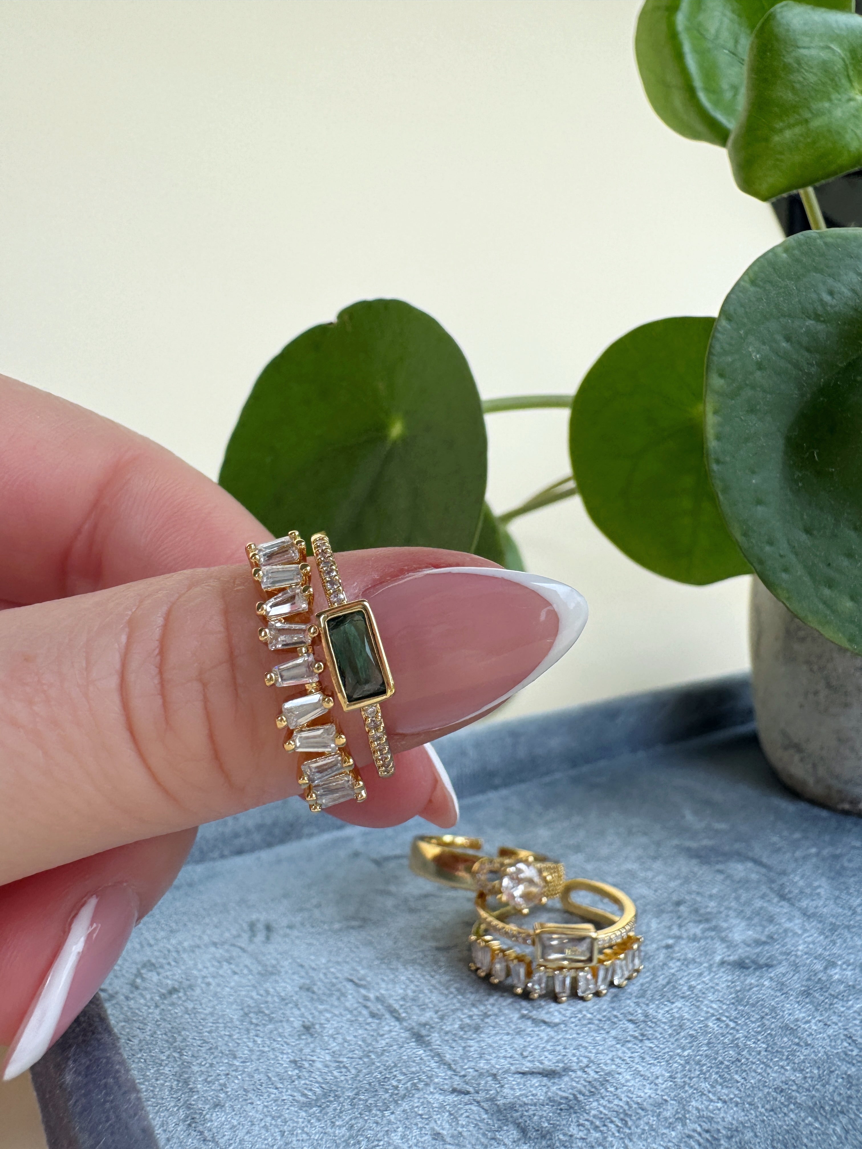 Sparkly gold ring with green gemstone held by a hand, with more rings on a blue surface and a plant in the background.
