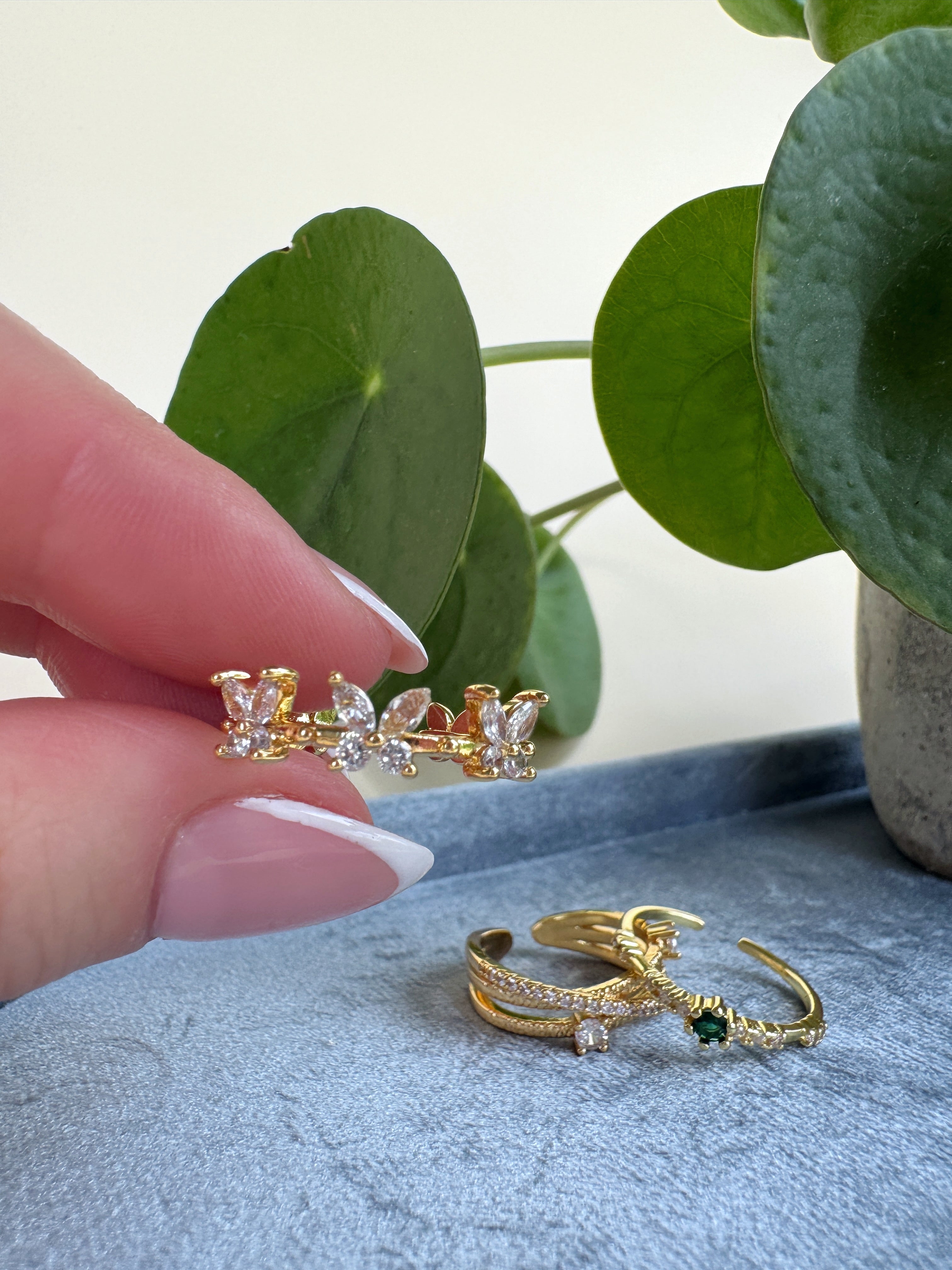 Gold butterfly ring with gemstones held by a classic french manicure hand against a light background with green leaves.