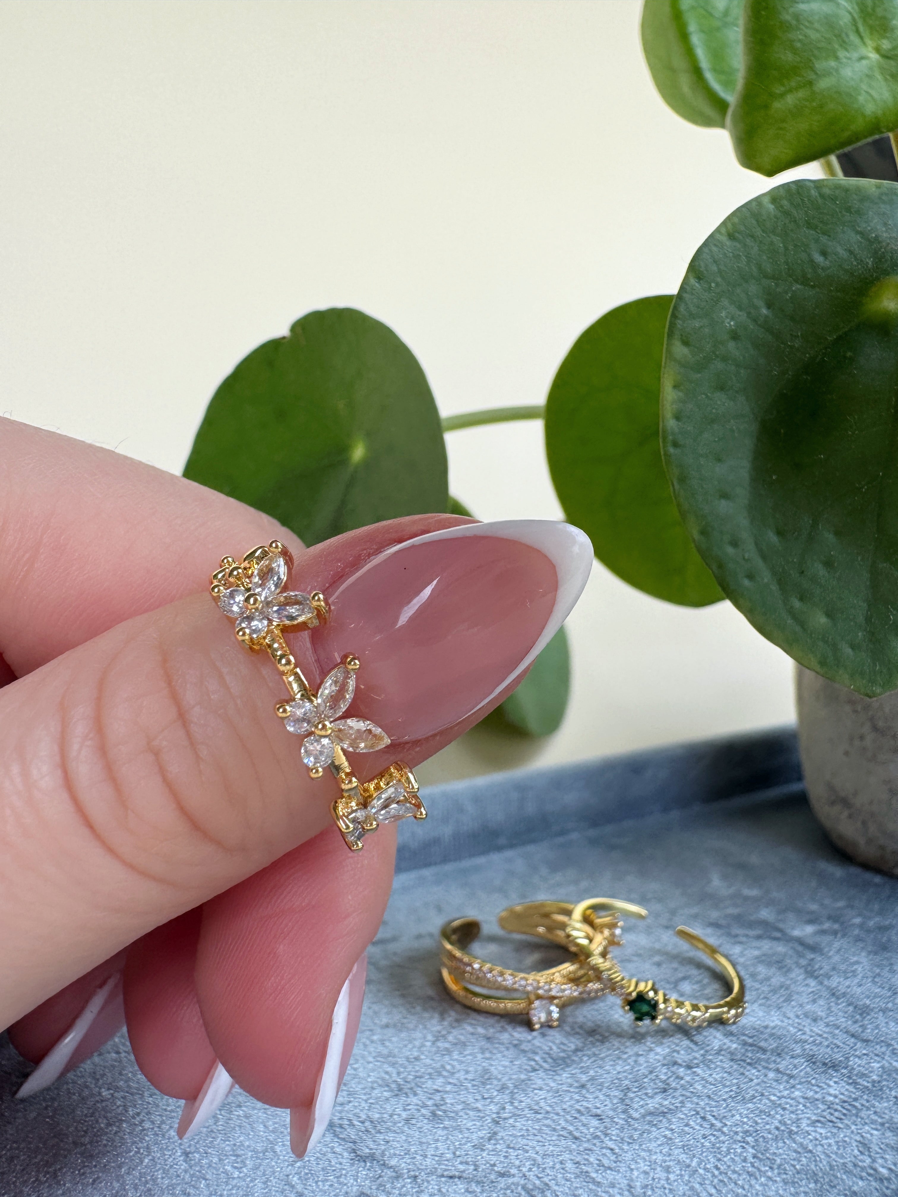 Gold butterfly ring held by a french manicured hand with a plant in the background