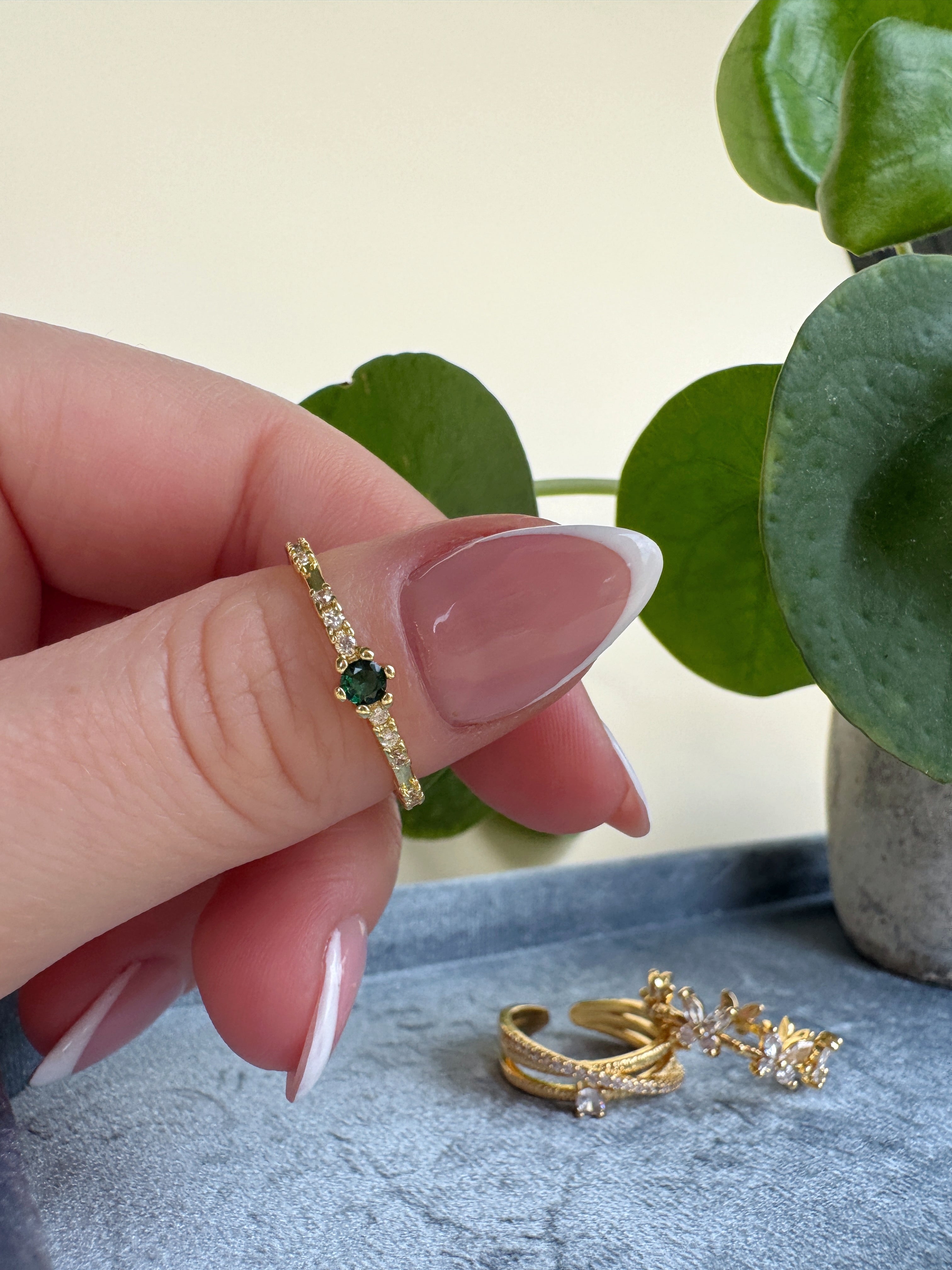 French manicure hand wearing a gold ring with a green gemstone, with additional gold rings on a textured surface.