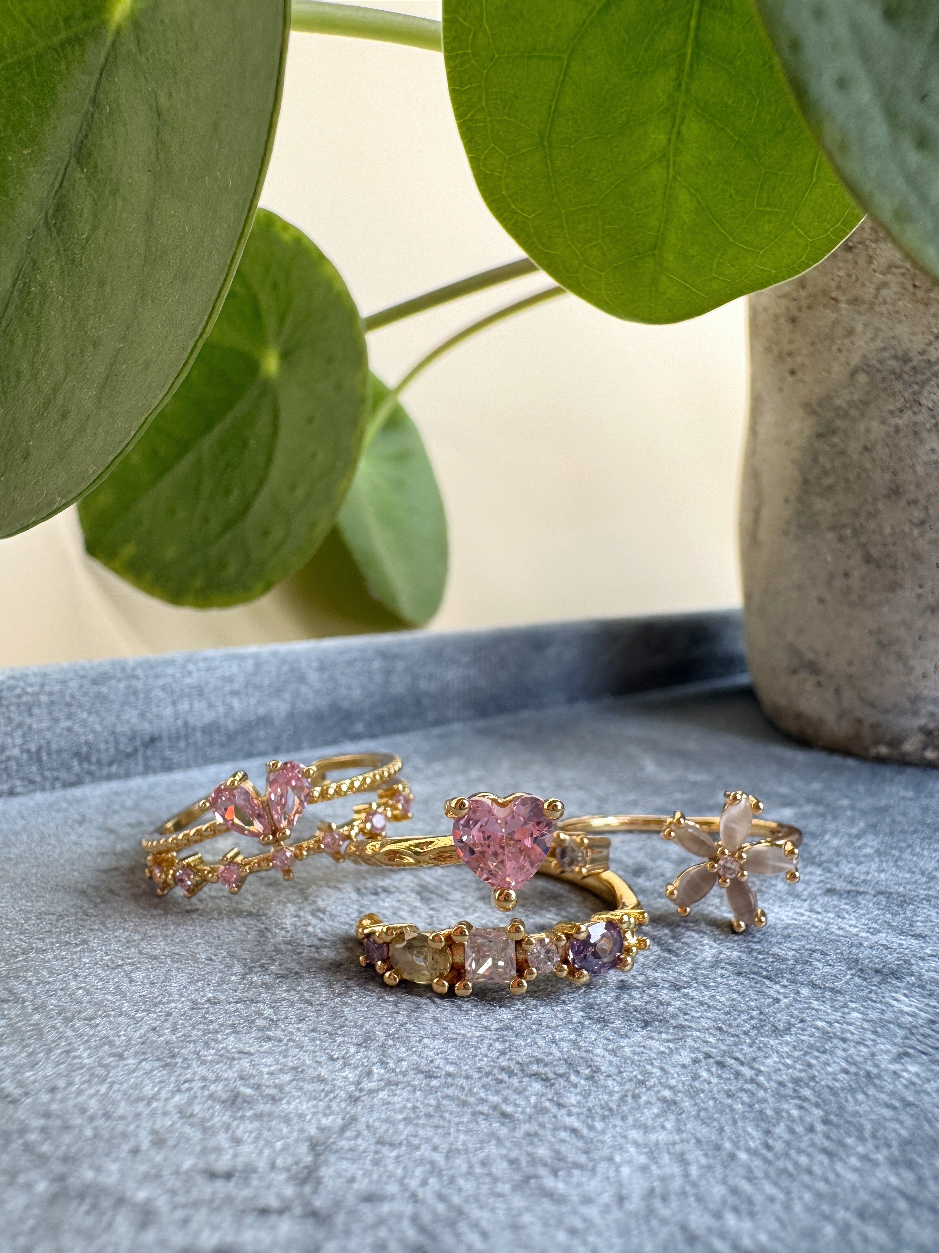 Set of dainty gold rings with pink and clear gemstones on a gray surface with green leaves in the background