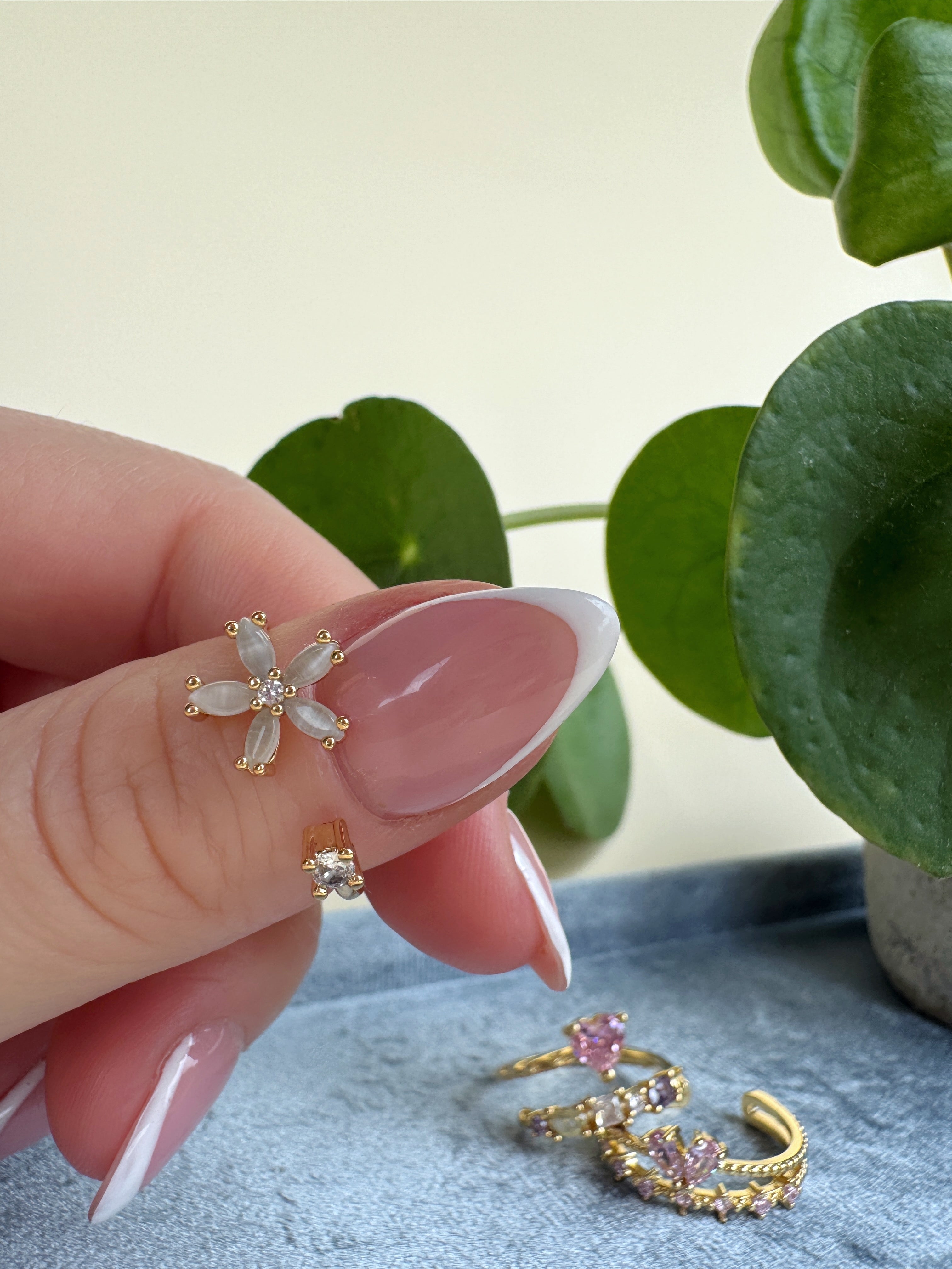 Hand holding a gold ring with a flower design, with more rings on a blue surface and green leaves in the background.