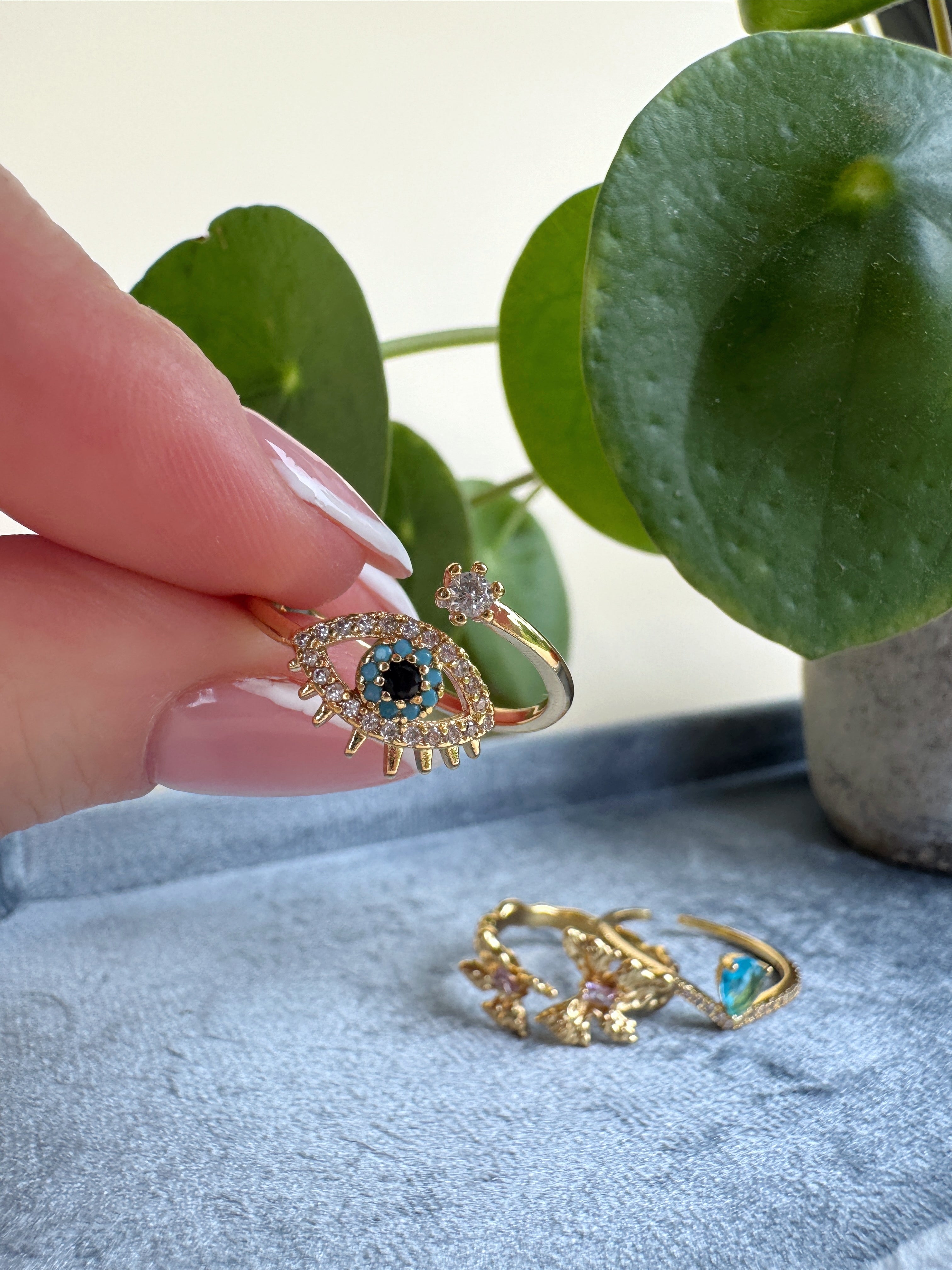 Gold evil eye ring with gemstones held by a hand, with another ring on a textured surface and a plant in the background.