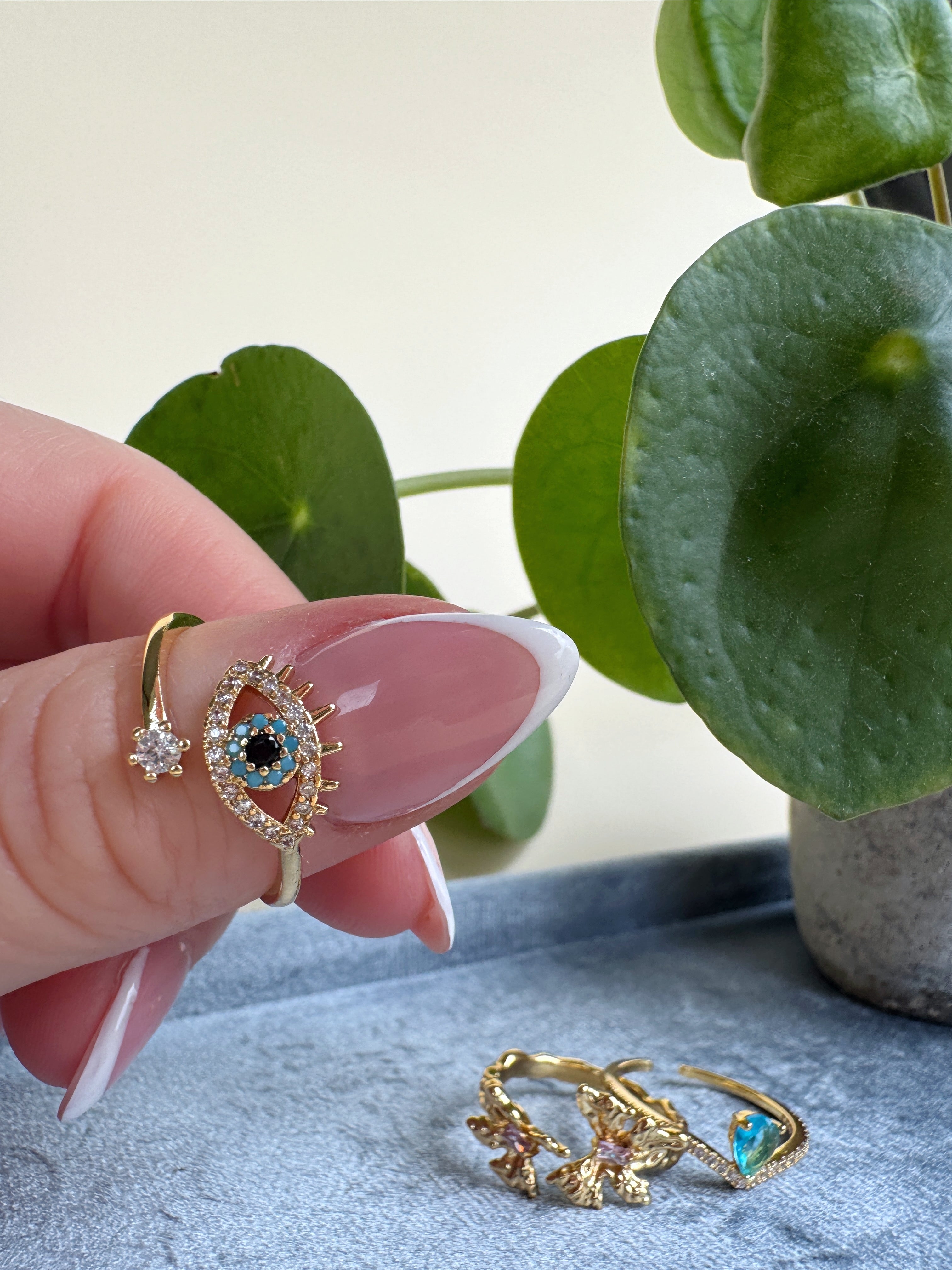 Gold evil eye ring with blue gemstone held by a hand against a light background with green leaves.