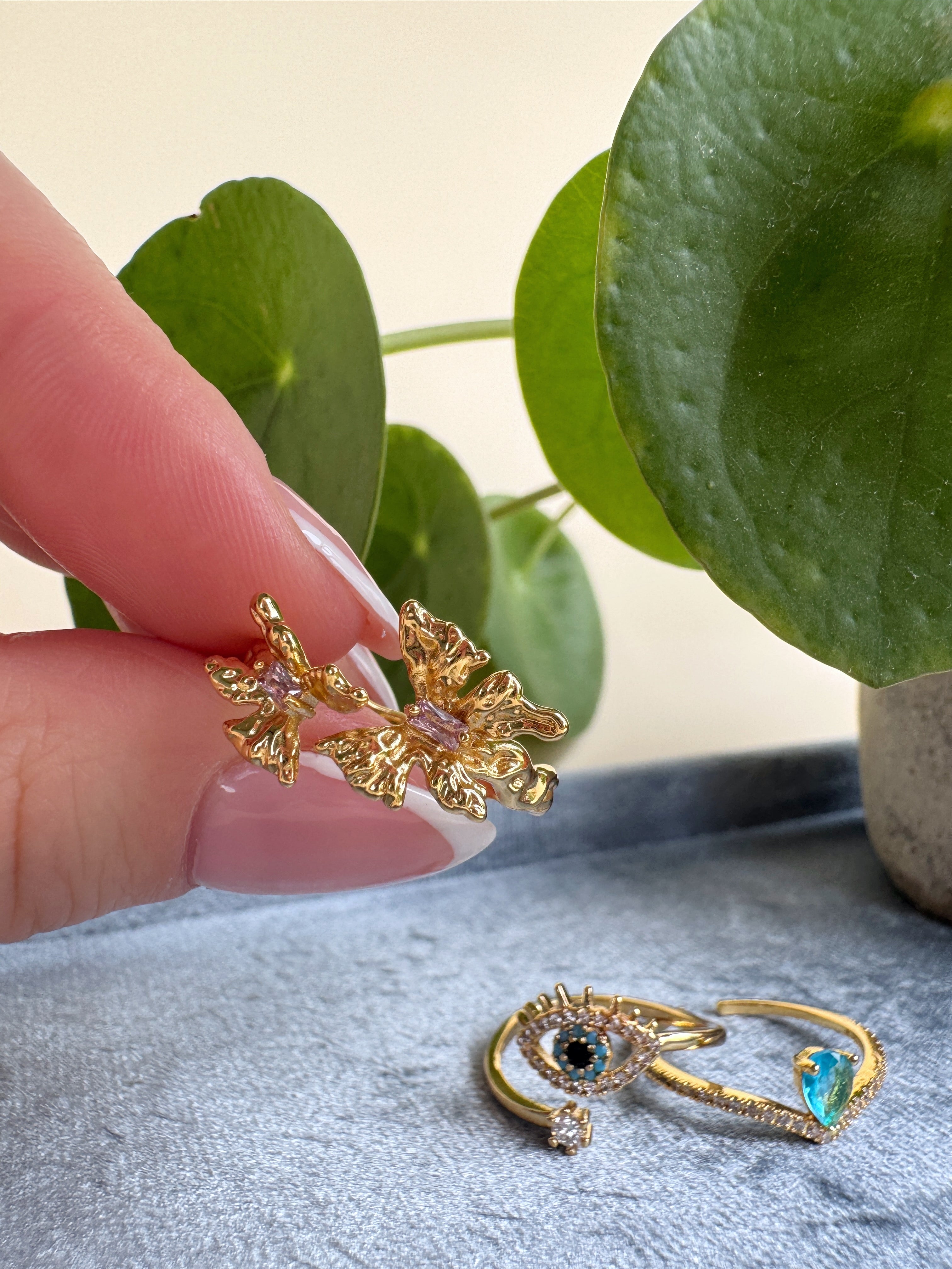 Gold ring with organic design held by a hand, with another ring featuring a blue gemstone on a textured surface.