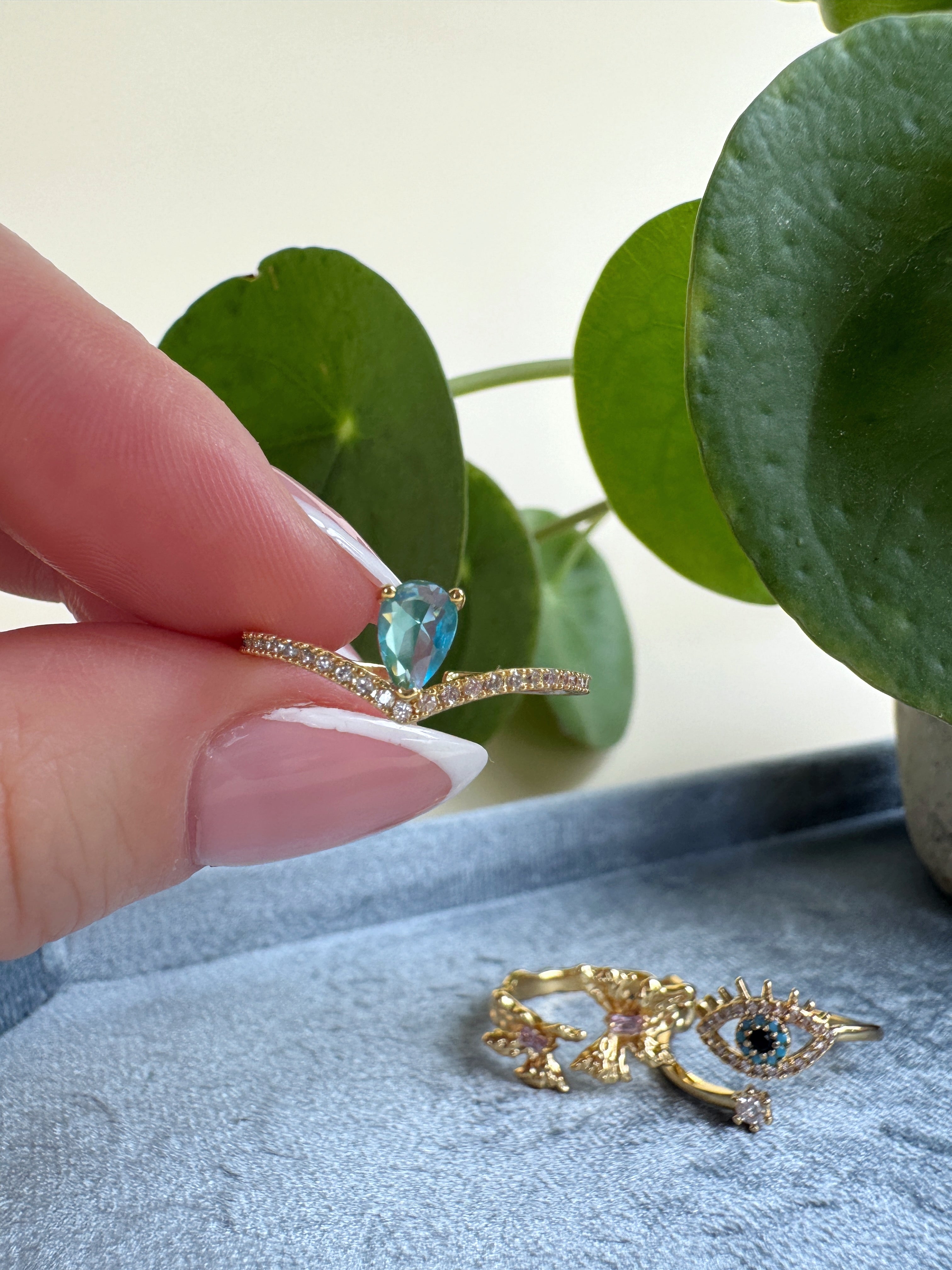 Feminine gold ring with a blue gemstone held by a hand, with another gold ring featuring an eye design on a textured surface.