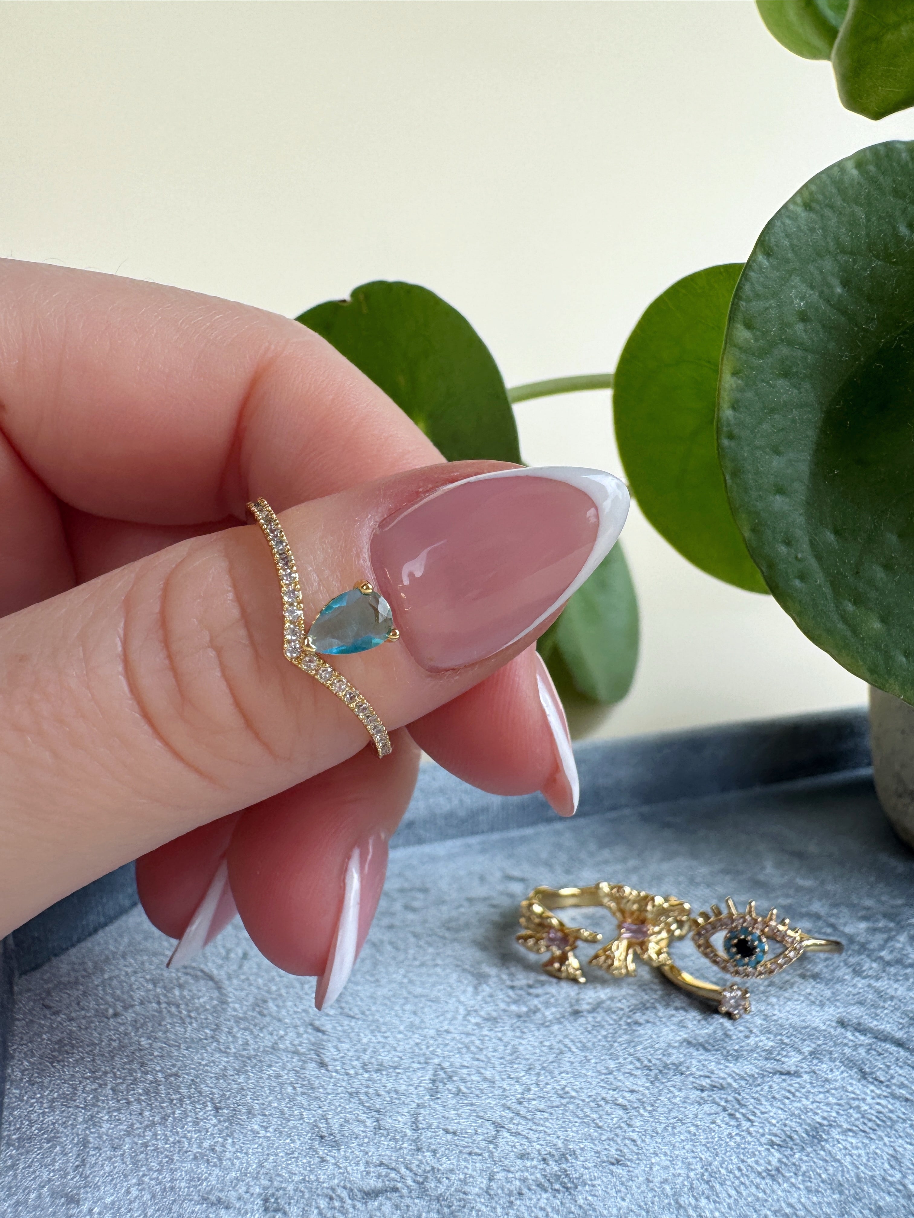 French manicure hand wearing a gold ring with a blue gemstone, with another gold ring on a surface in the background.