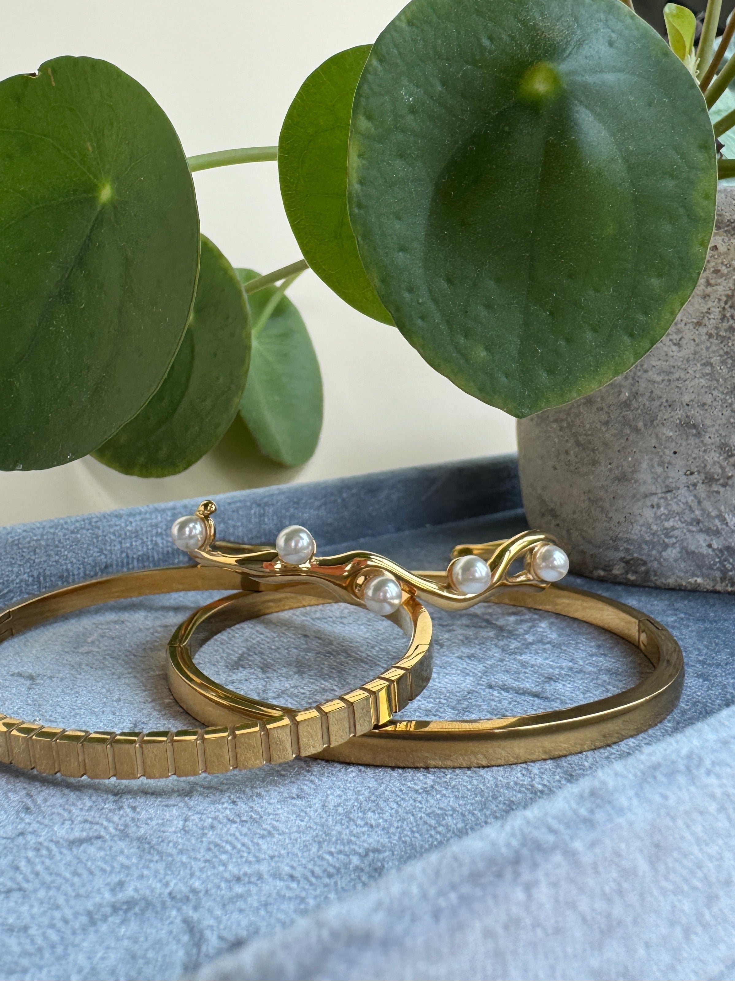 Set of 3 gold bracelets with pearl accents on a blue surface next to a green plant.