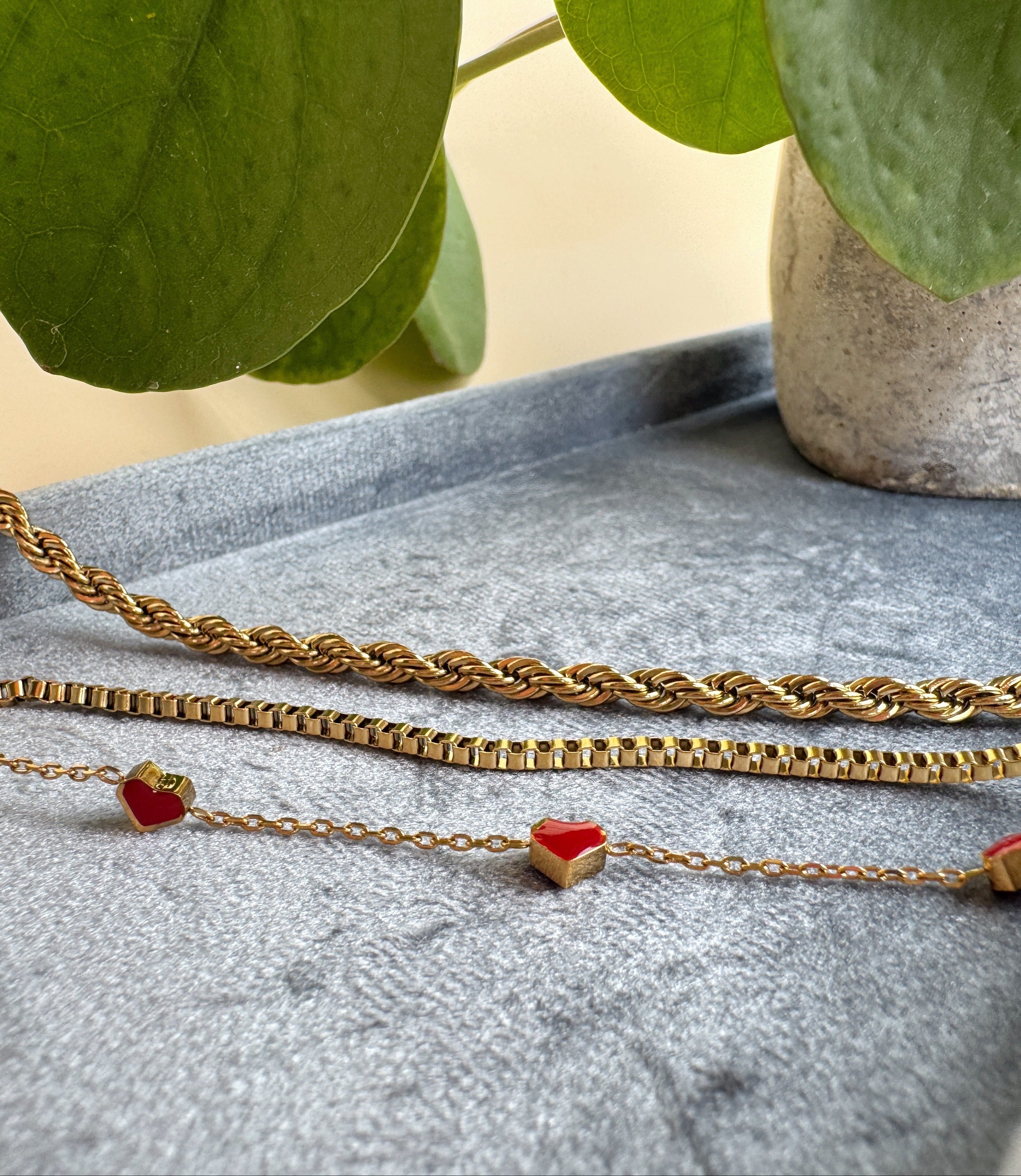 Gold chain necklace with red heart pendants on a gray surface with green leaves in the background