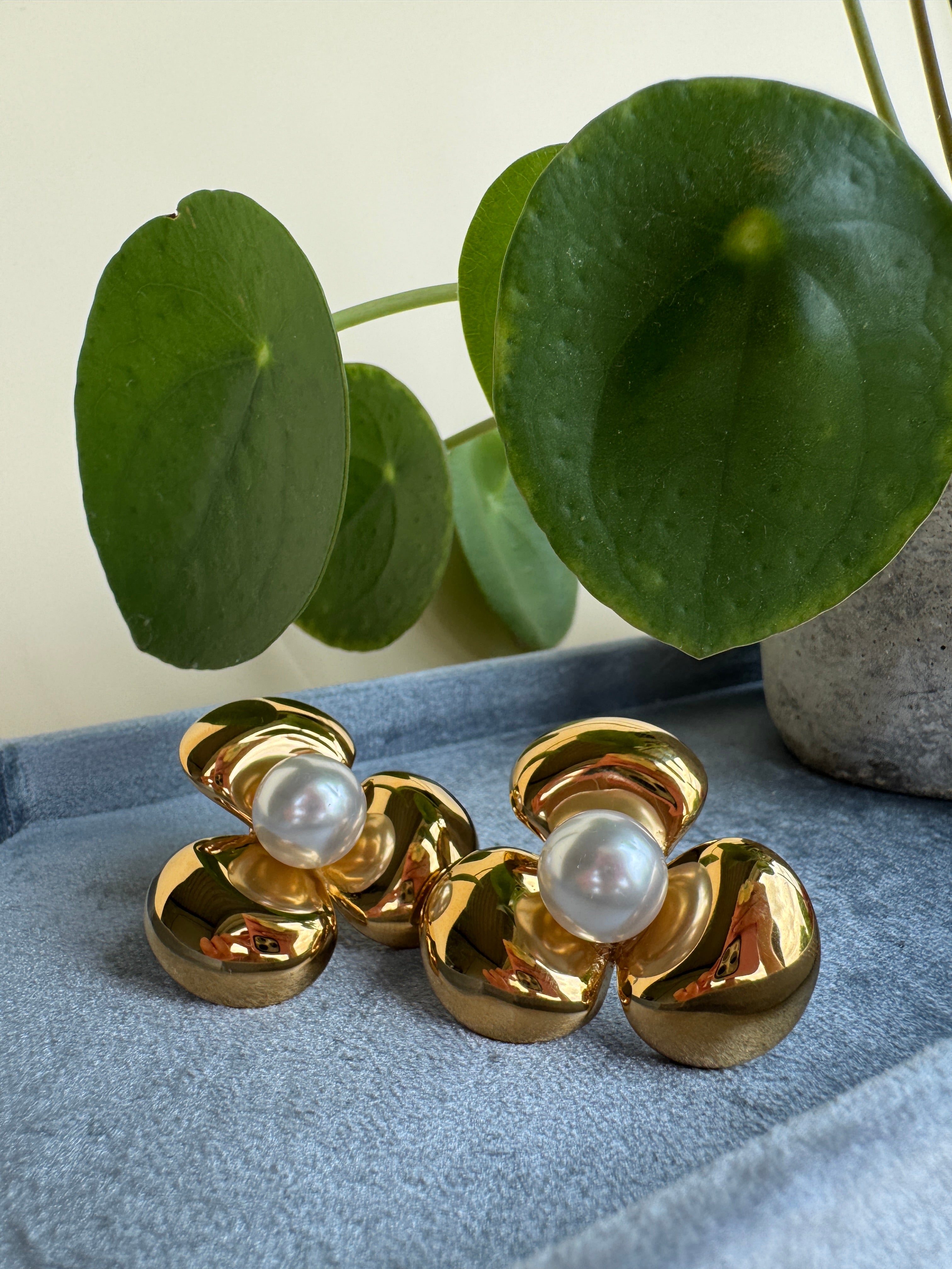 Gold flower earrings with pearls on a gray surface next to green leaves