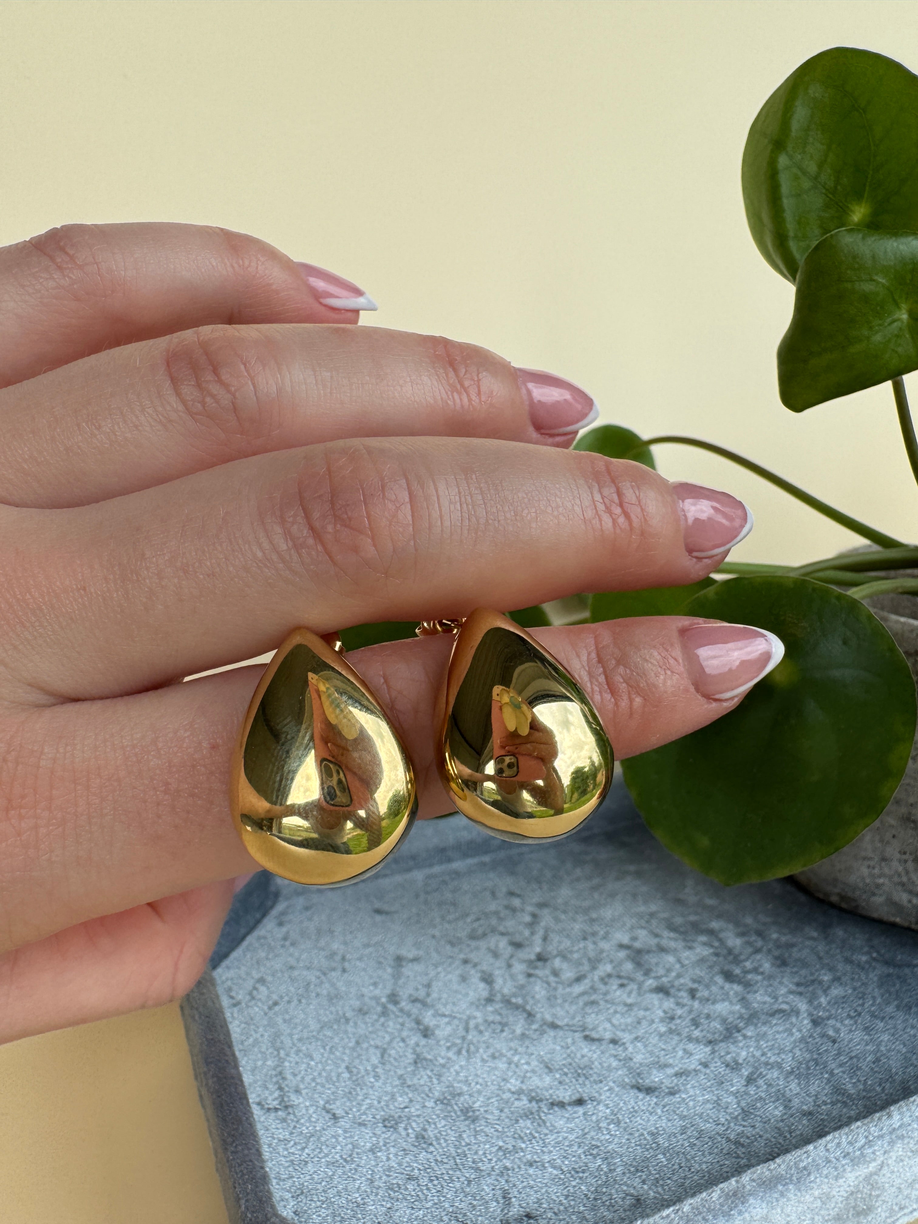 Gold teardrop earrings worn on a hand with green leaves in the background. Waterproof stainless steel.