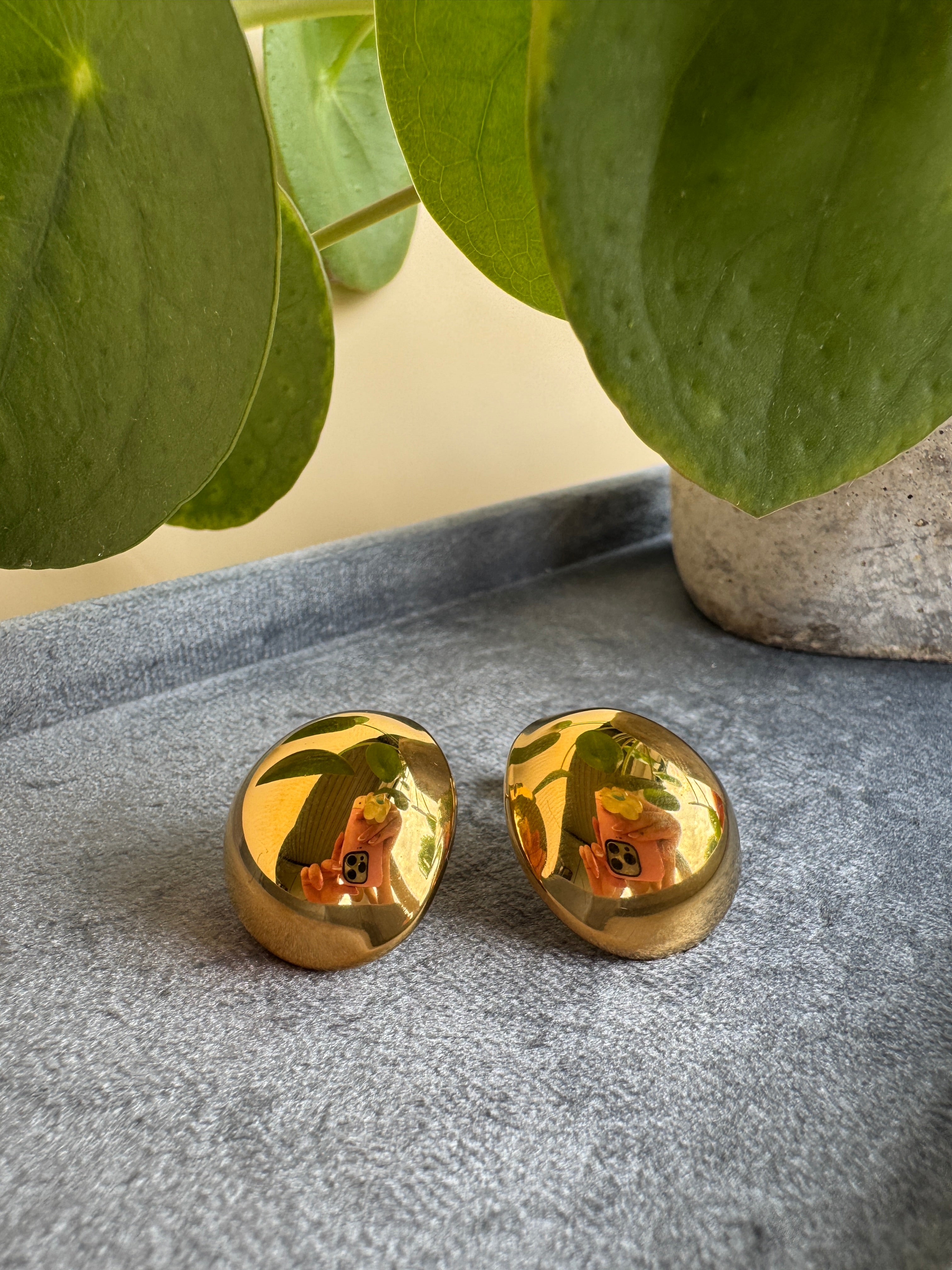 Two gold waterproof earrings on a textured surface with green leaves in the background