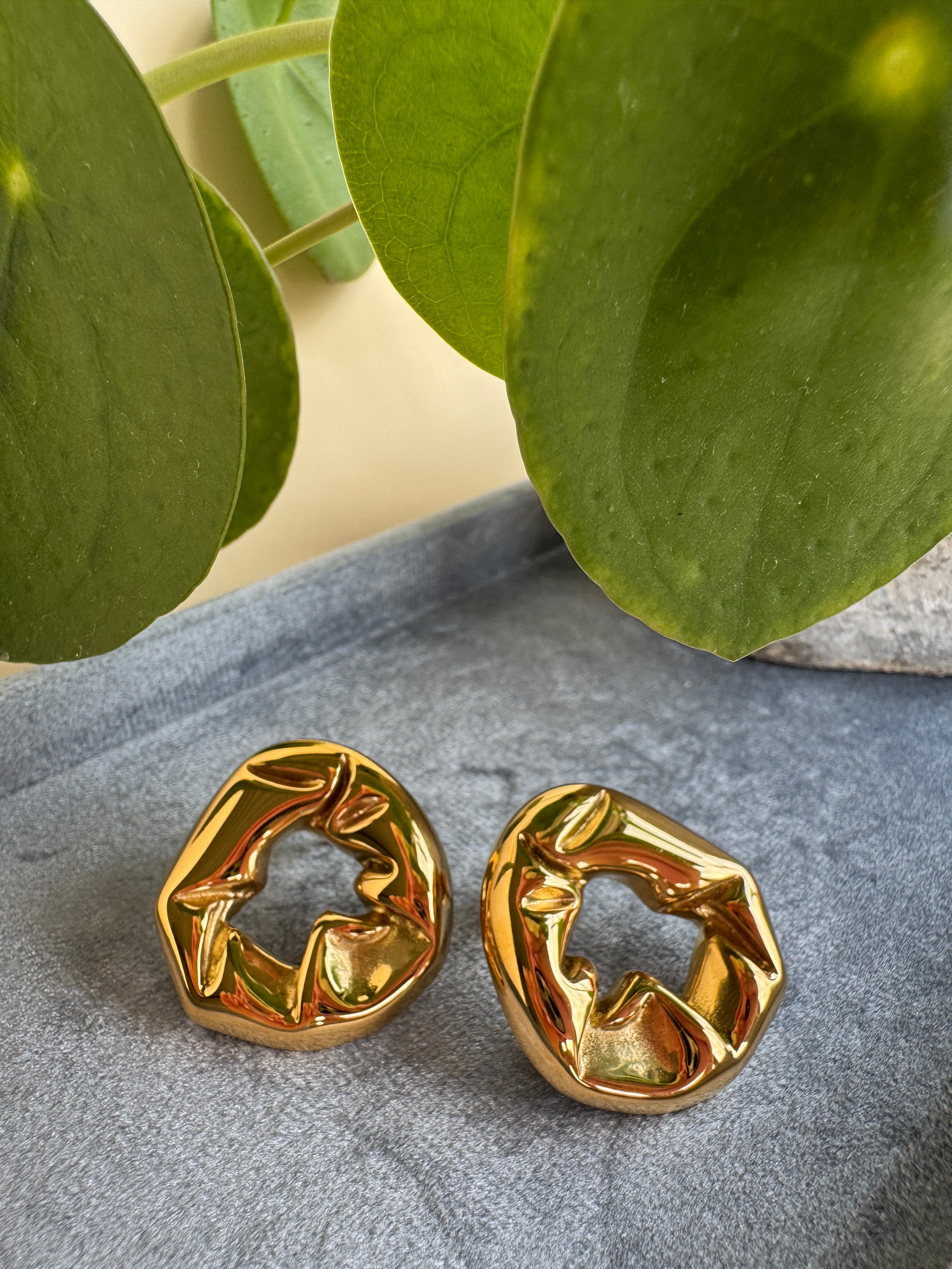 Unique gold earrings on a textured surface with green leaves in the background