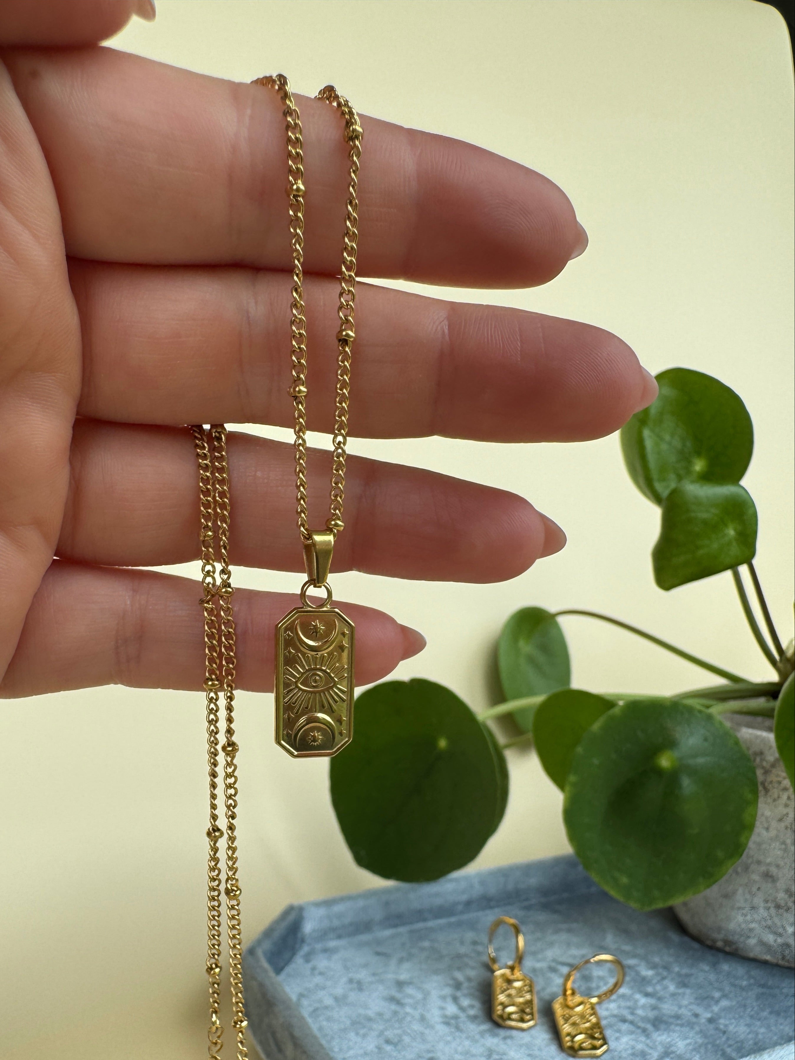 Gold evil eye necklace on a hand with a plant in the background
