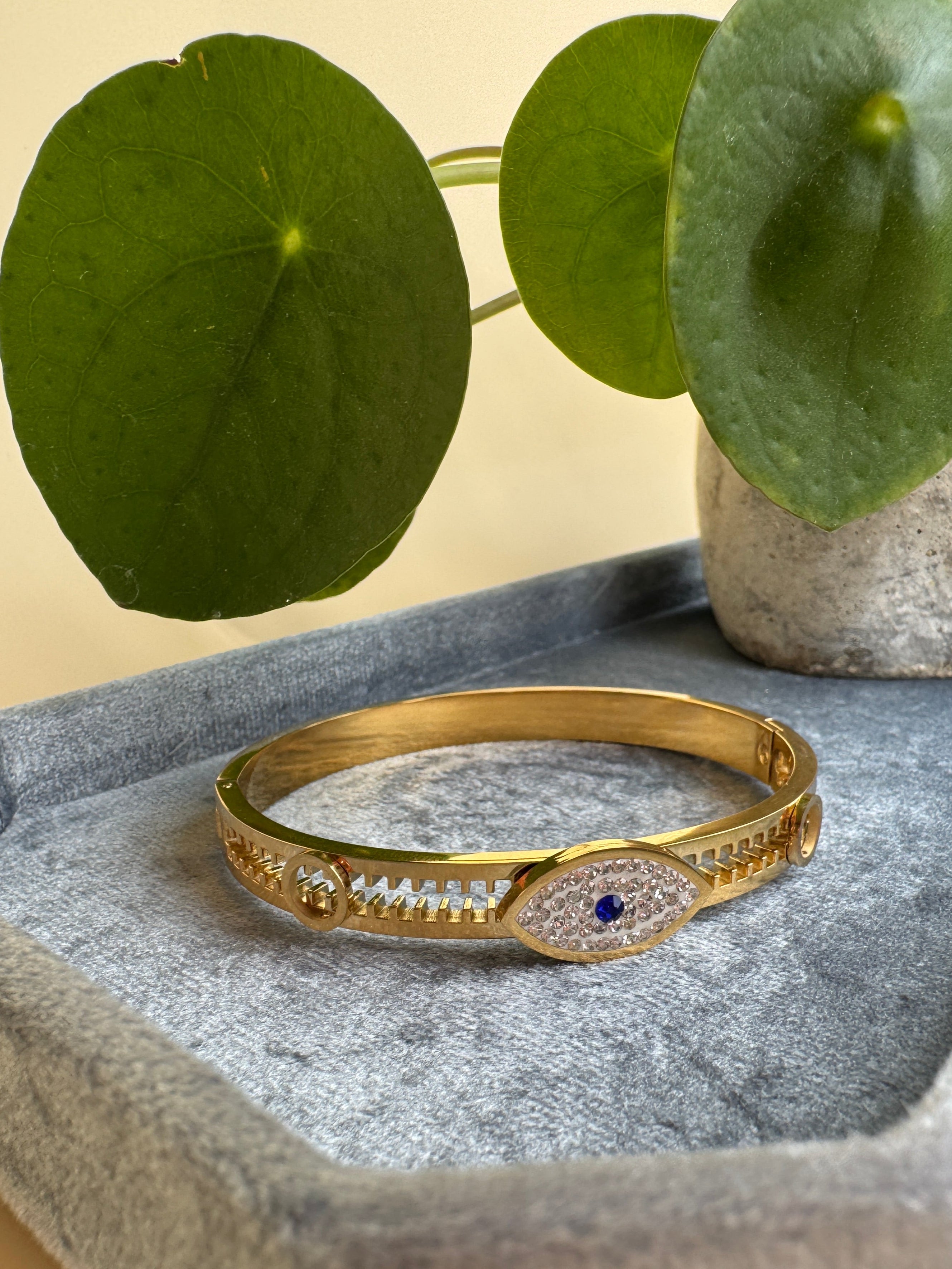 Gold evil eye good luck bracelet with a blue gemstone on a textured surface with green leaves in the background
