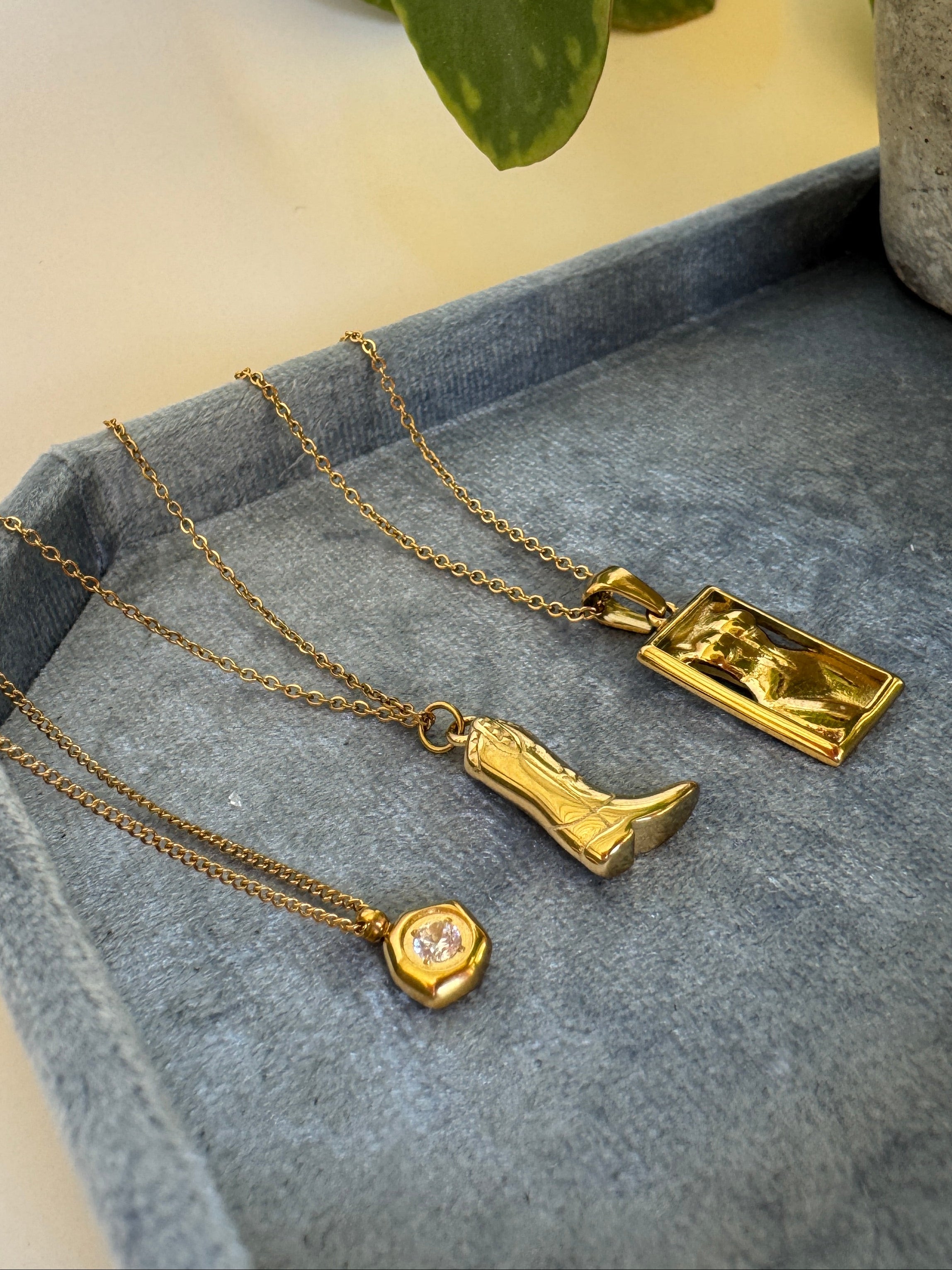 Three gold necklaces with pendants on a gray surface, with a plant in the background.