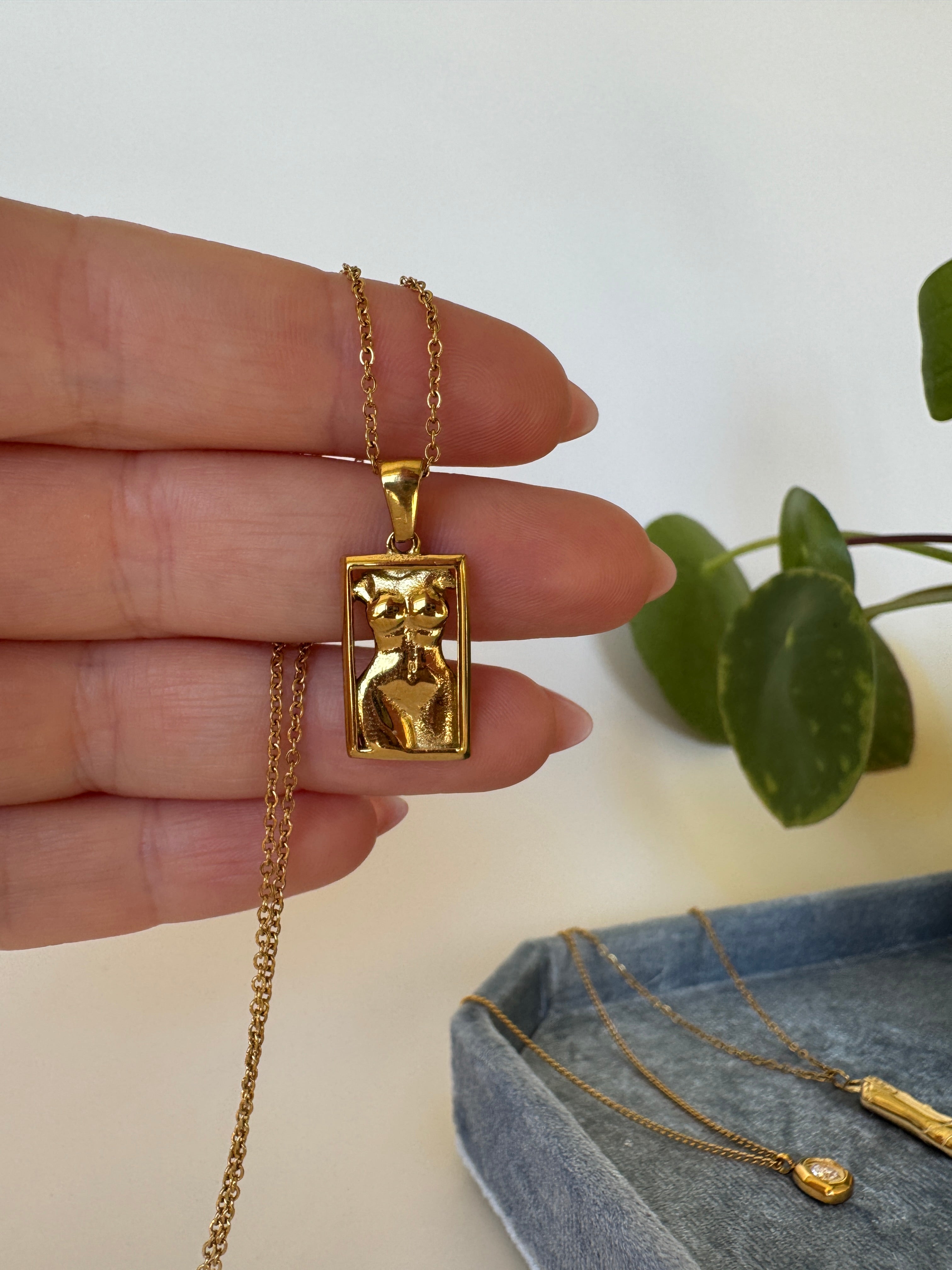 Gold necklace with a woman torso / figure pendant held by a hand against a neutral background