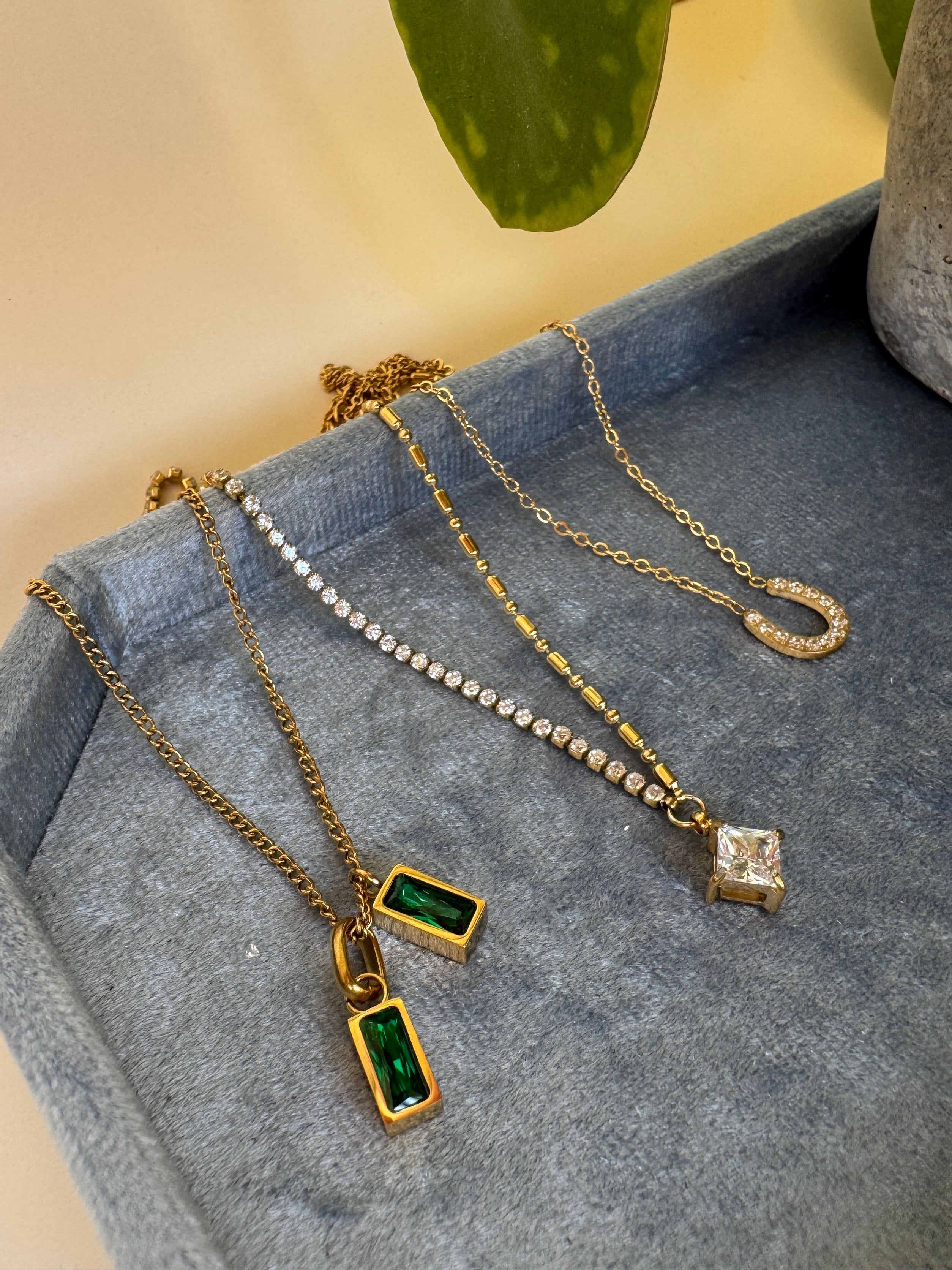 Two gold necklaces with green and clear pendants on a denim surface with a blurred plant background.