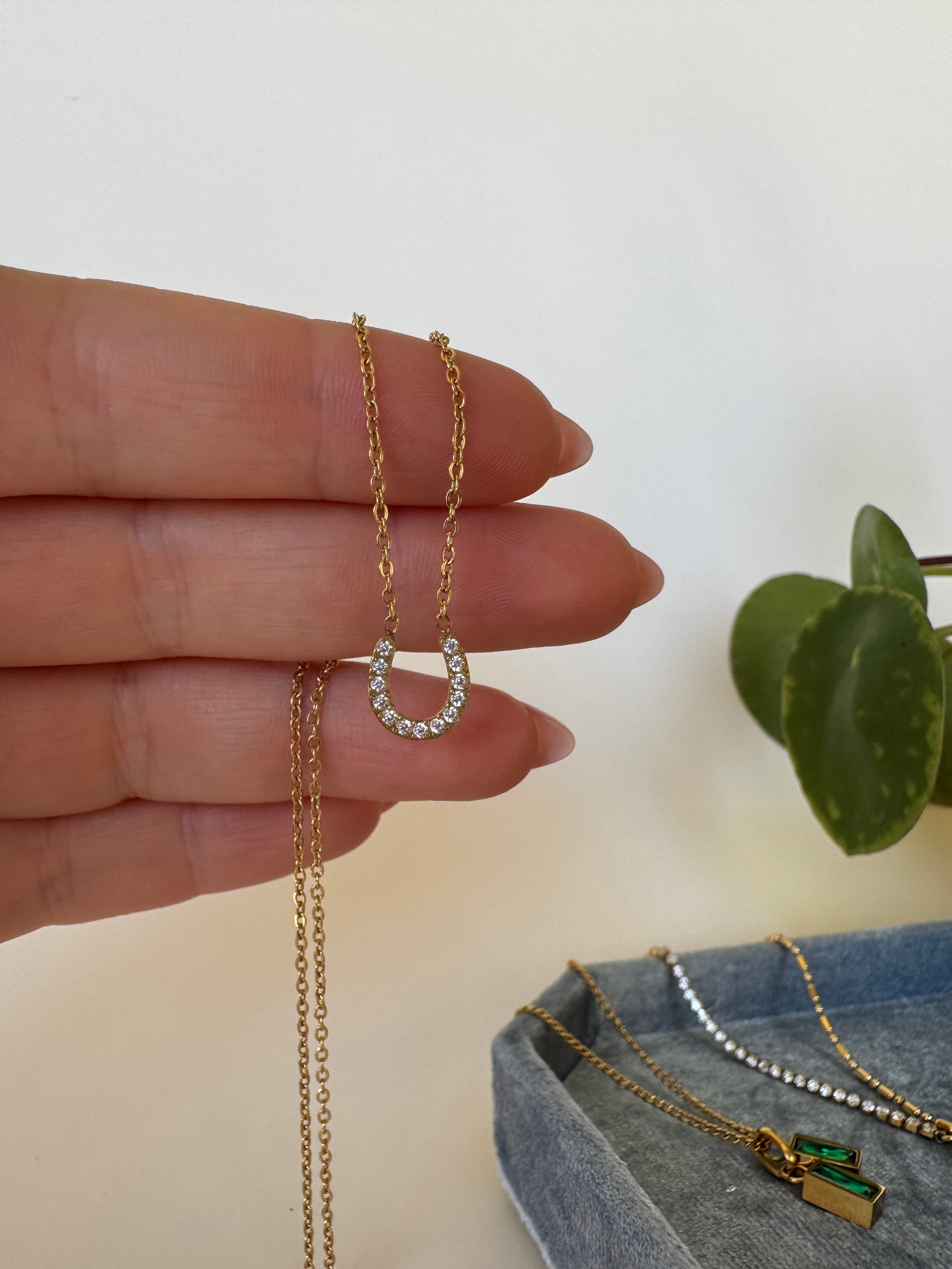 Close-up of a hand wearing a gold horse shoe necklace with a dainty pendant on a neutral background.