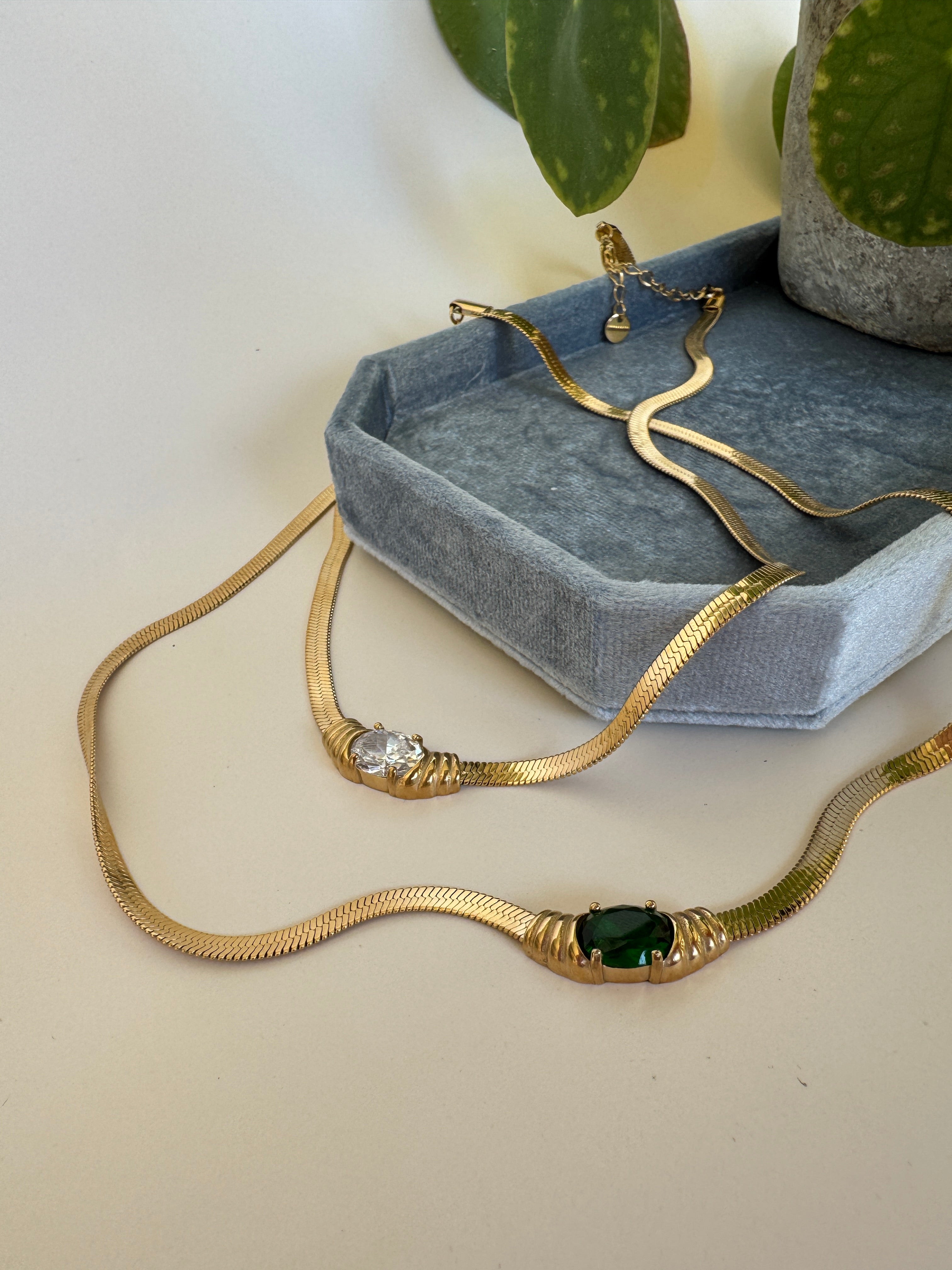 Gold necklaces with green and white gemstones on a gray cushion with a plant in the background