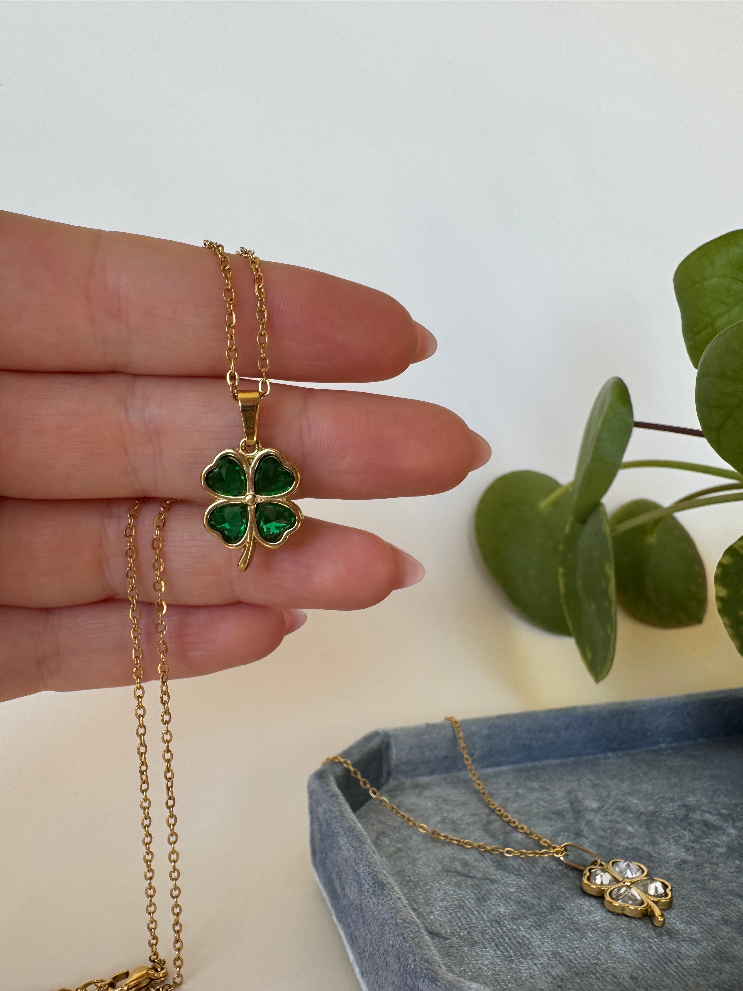 Gold ring with green four leaf clover gemstone on a finger, with a necklace and plant in the background.