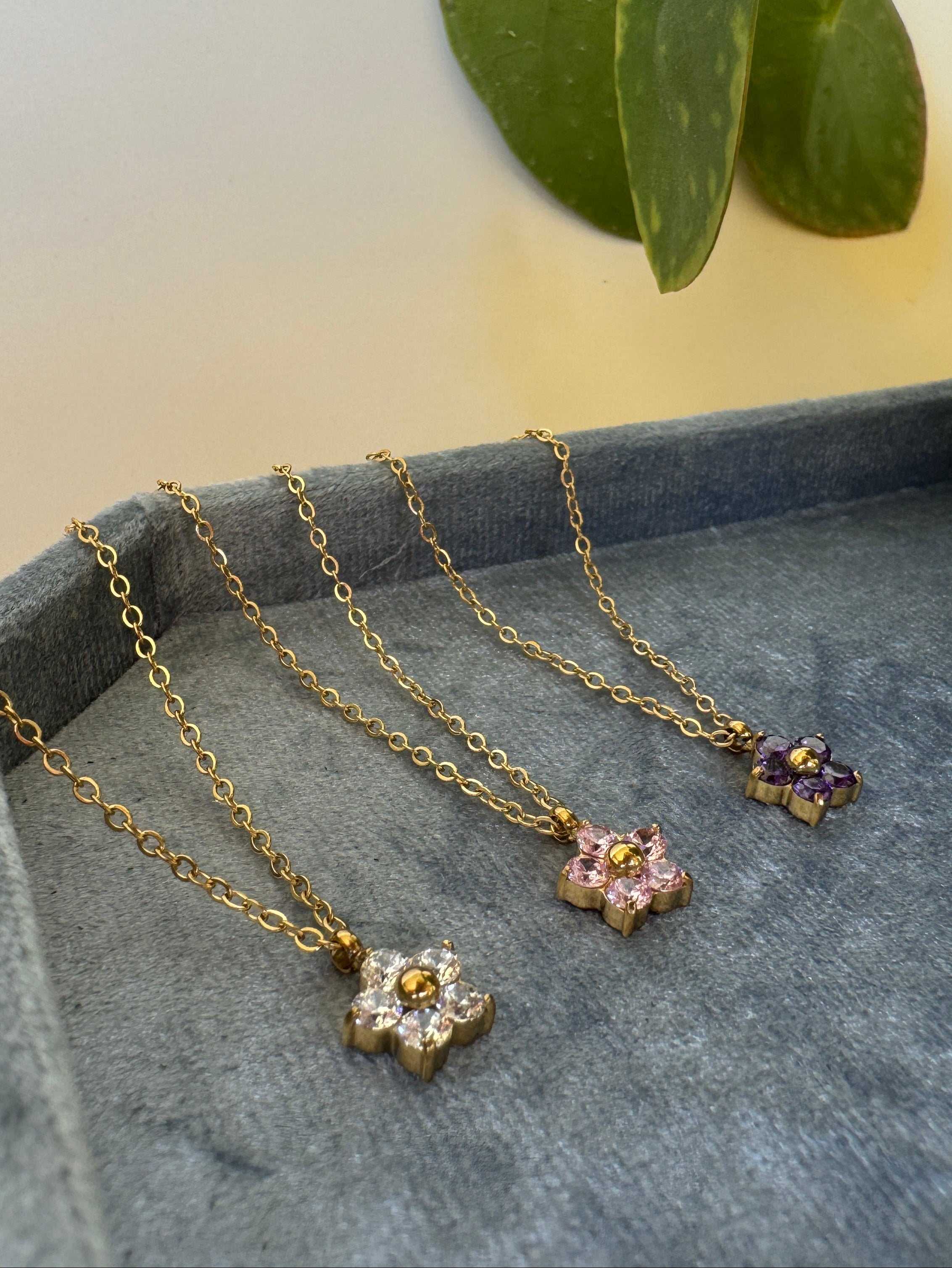 Gold stainless steel necklaces with floral pendants on a gray surface with a plant in the background