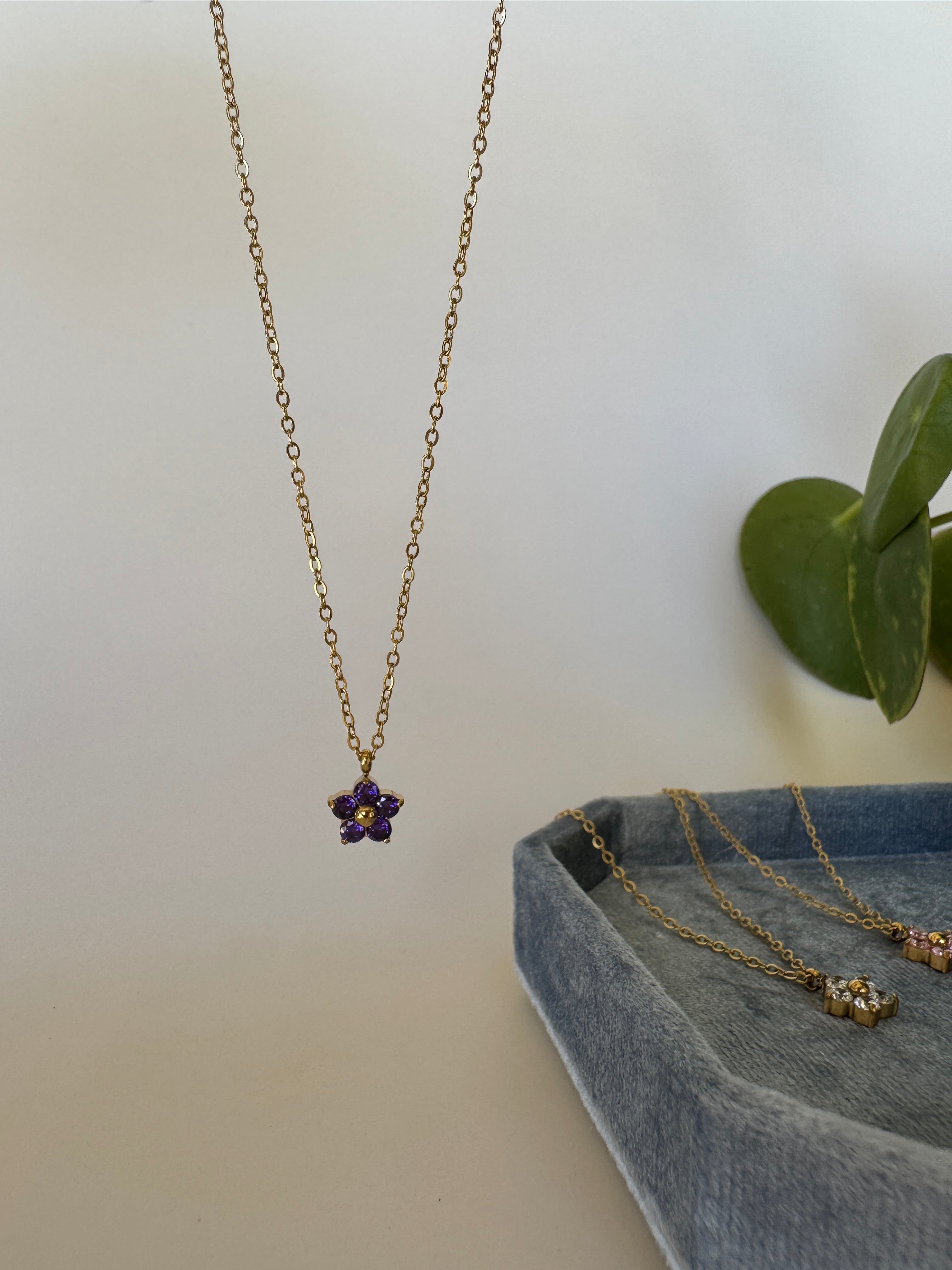Gold necklace with a purple gemstone on a white surface with a gray fabric and green leaves.