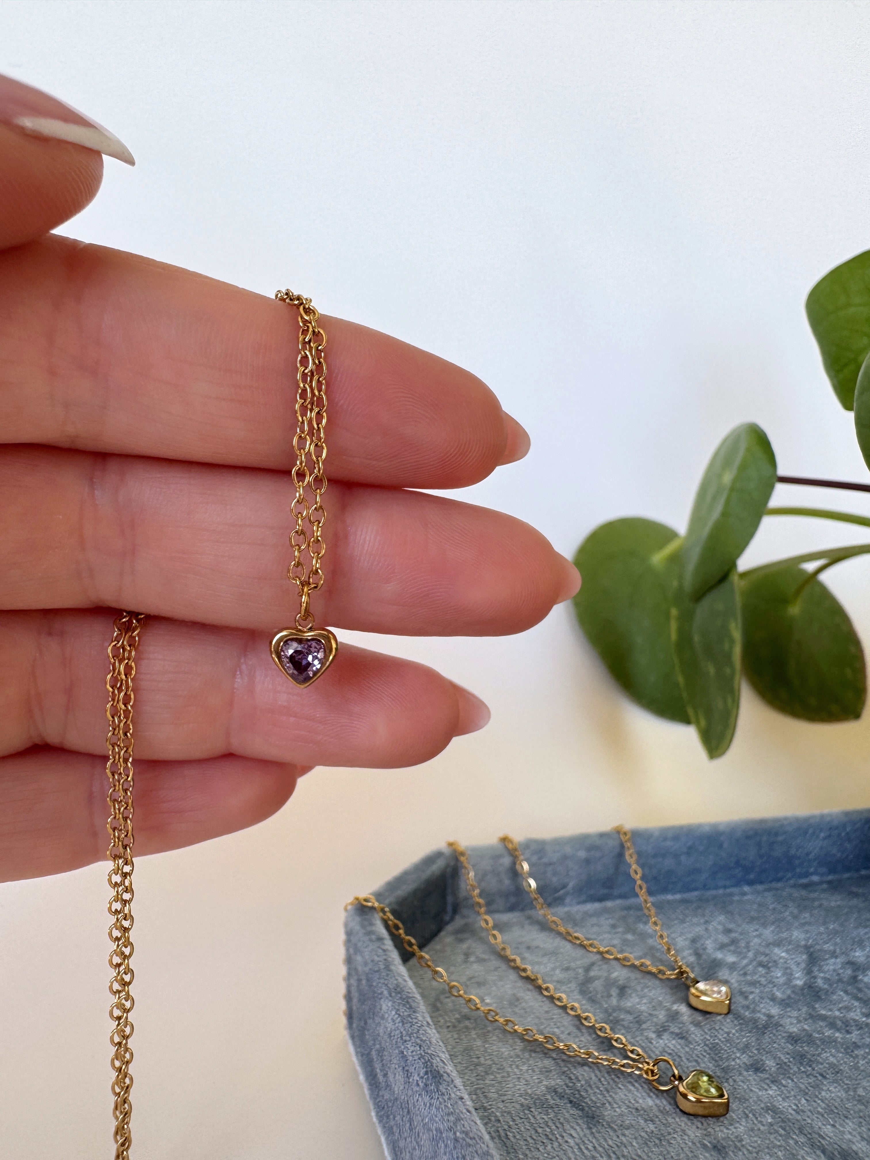 Gold heart-shaped necklace with a purple gemstone held by a hand, with a plant in the background.