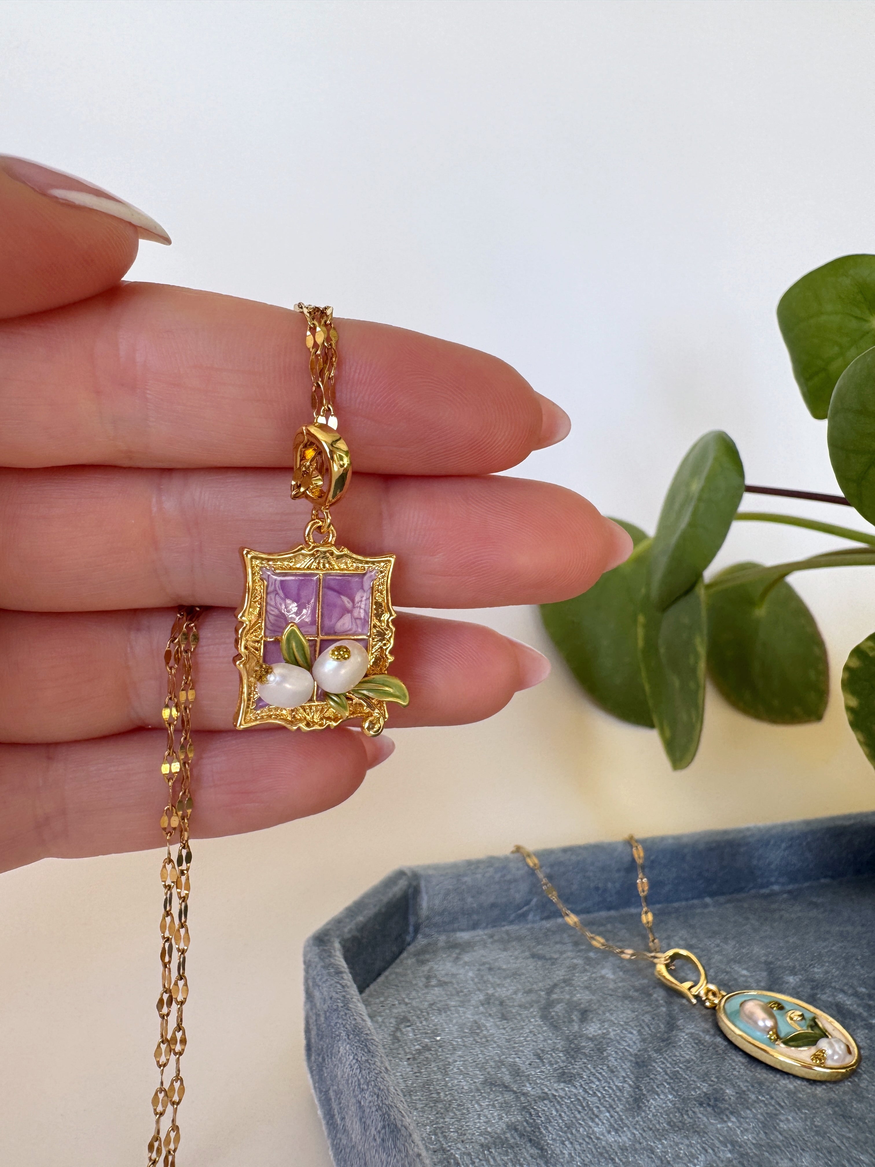 Gold necklace with a purple pendant held by a hand, with a plant in the background.