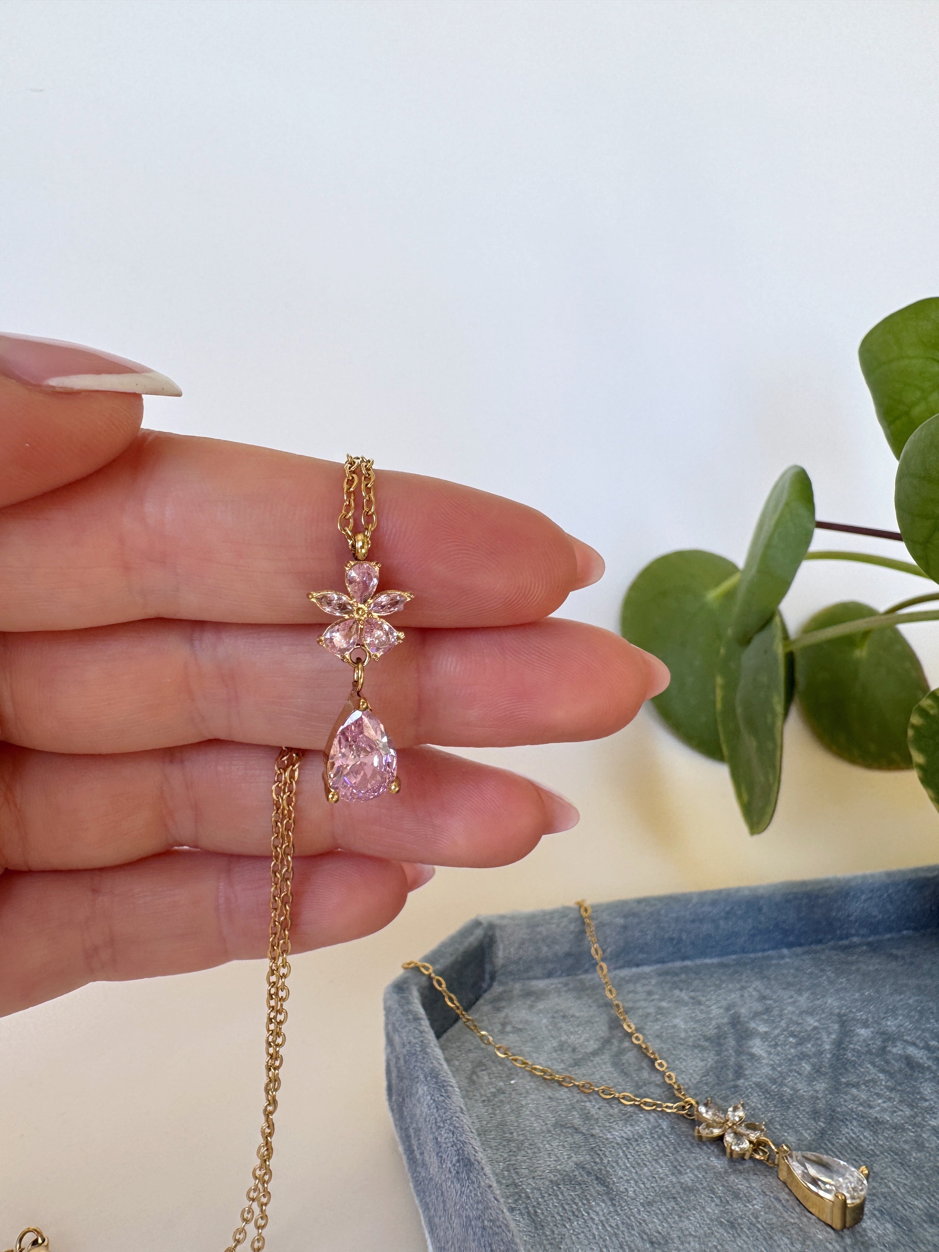 Hand holding a gold necklace with a pink teardrop pendant against a light background