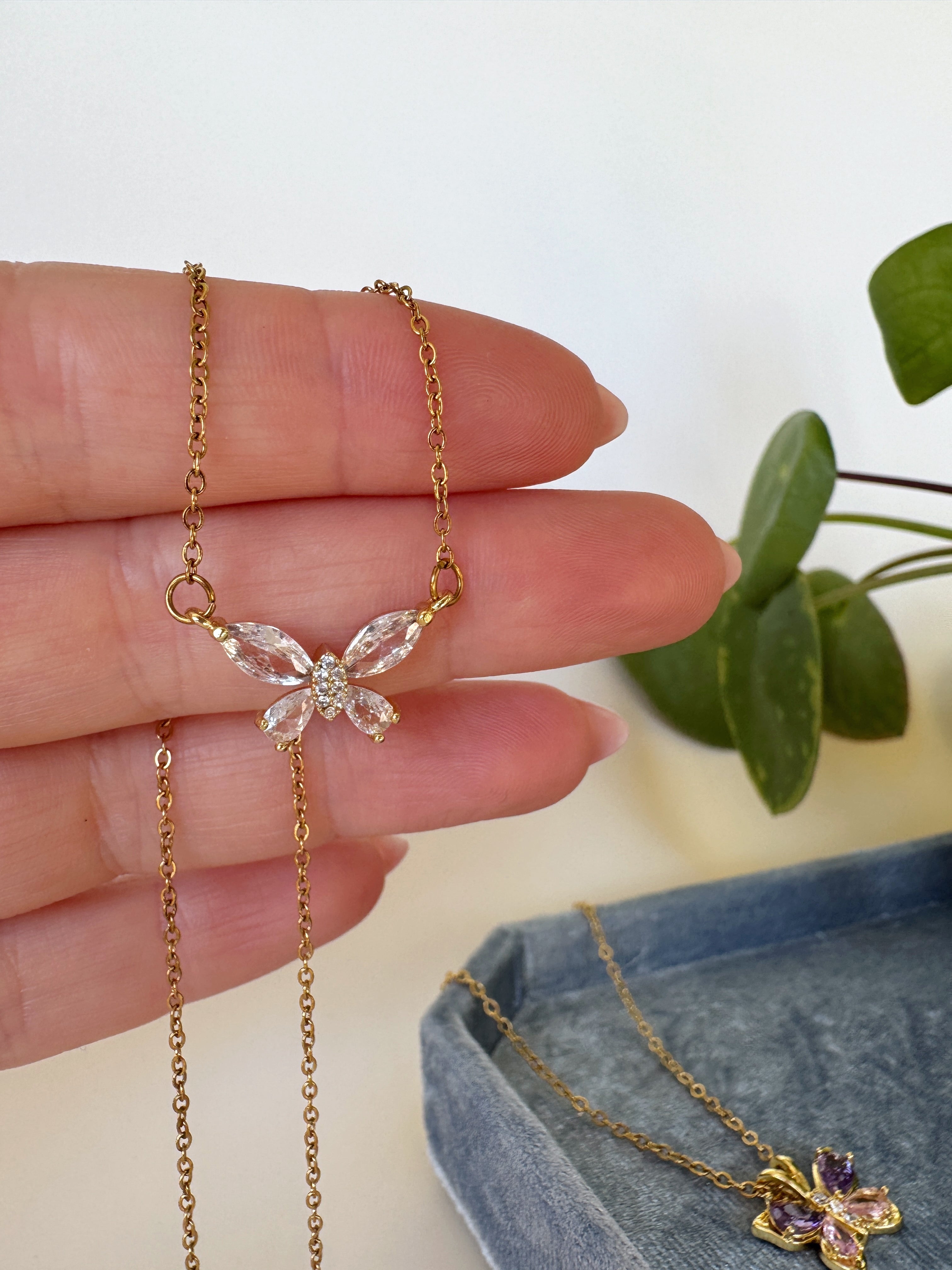 Gold butterfly necklace held by a hand with a plant in the background