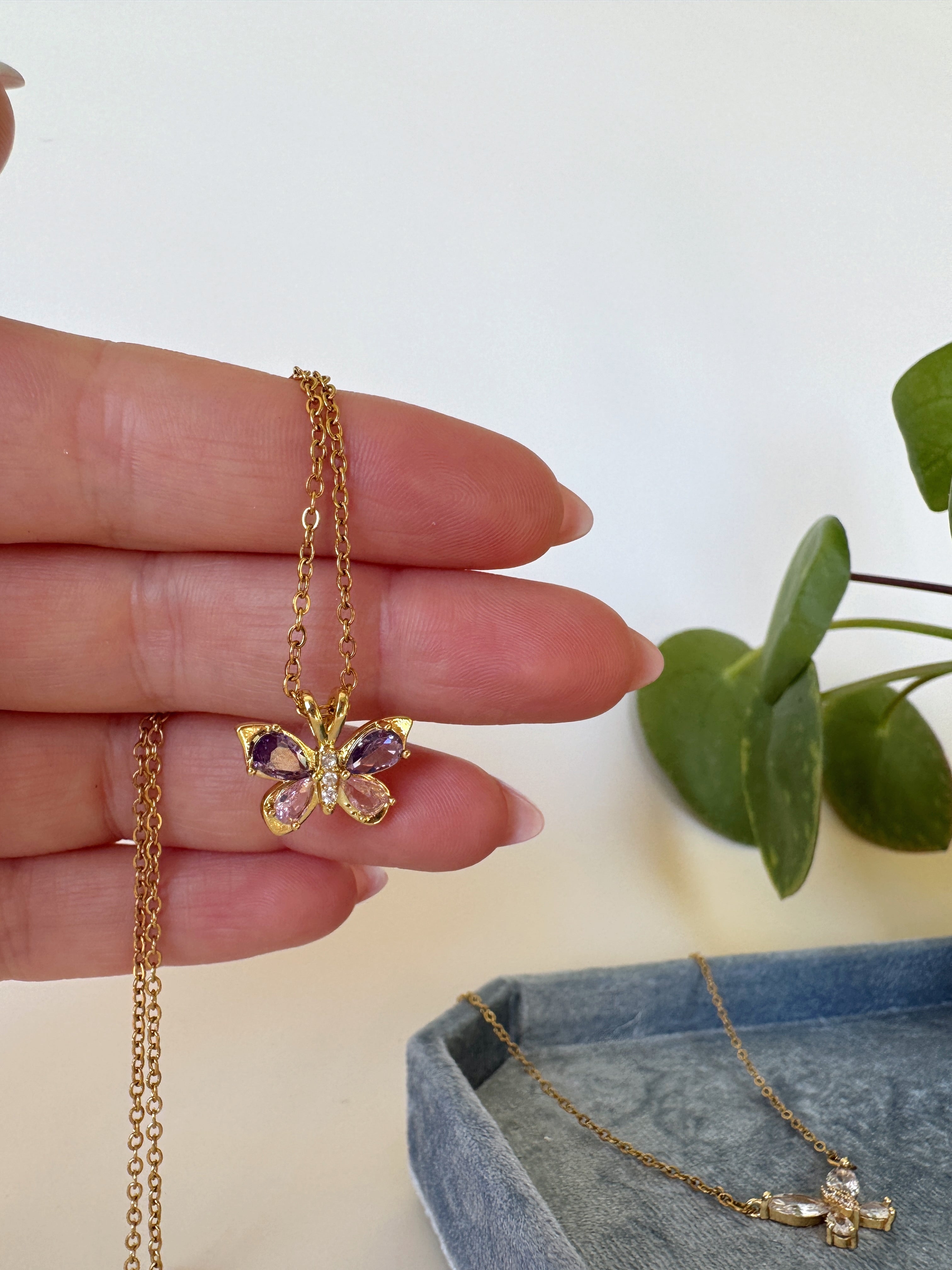 Butterfly-shaped gold necklace with gemstones held by a hand, with a plant in the background.