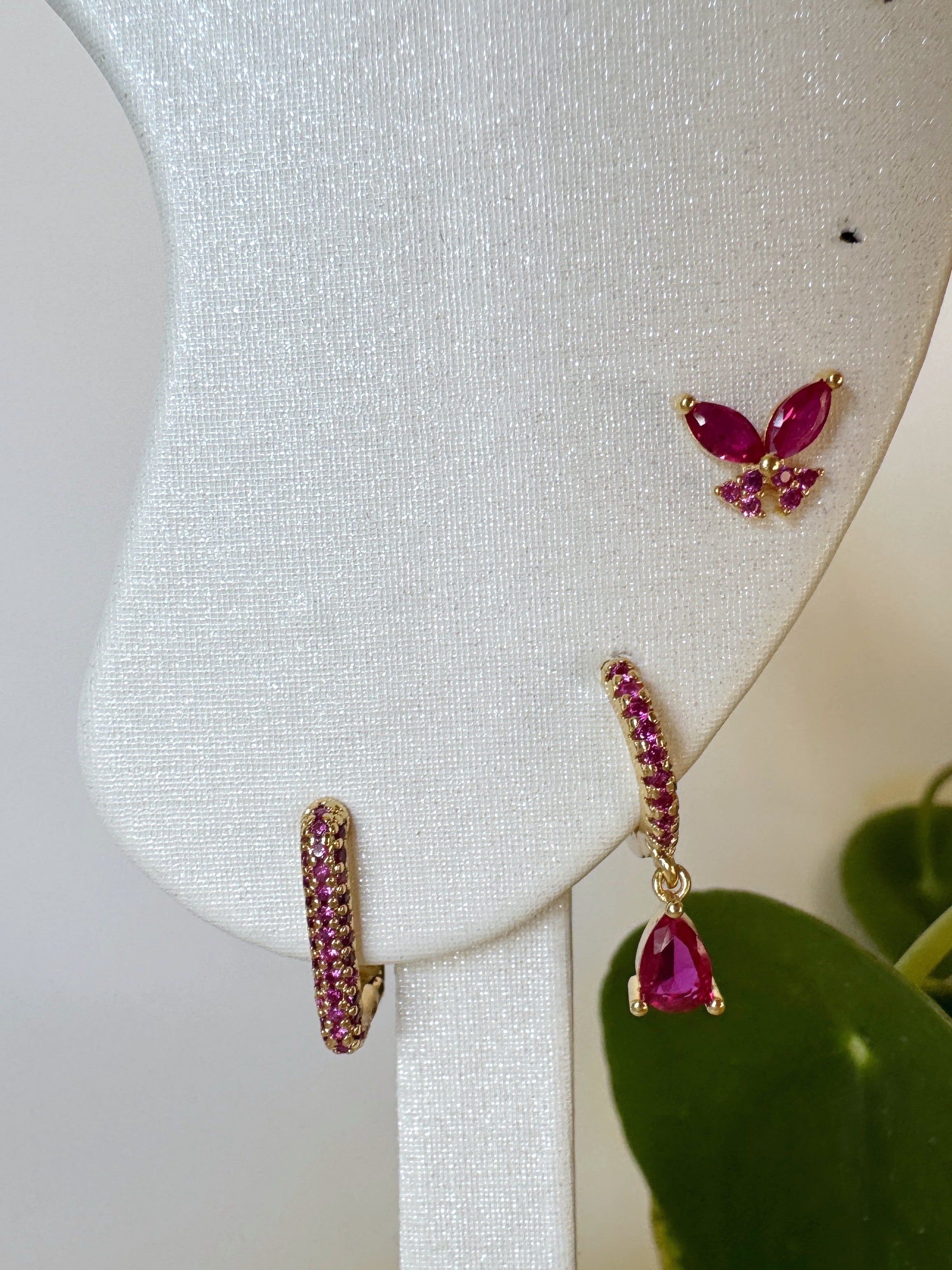 Butterfly-shaped waterproof stud earrings with dark pink gemstones on a white stand. Square hoops and dangly hoops.