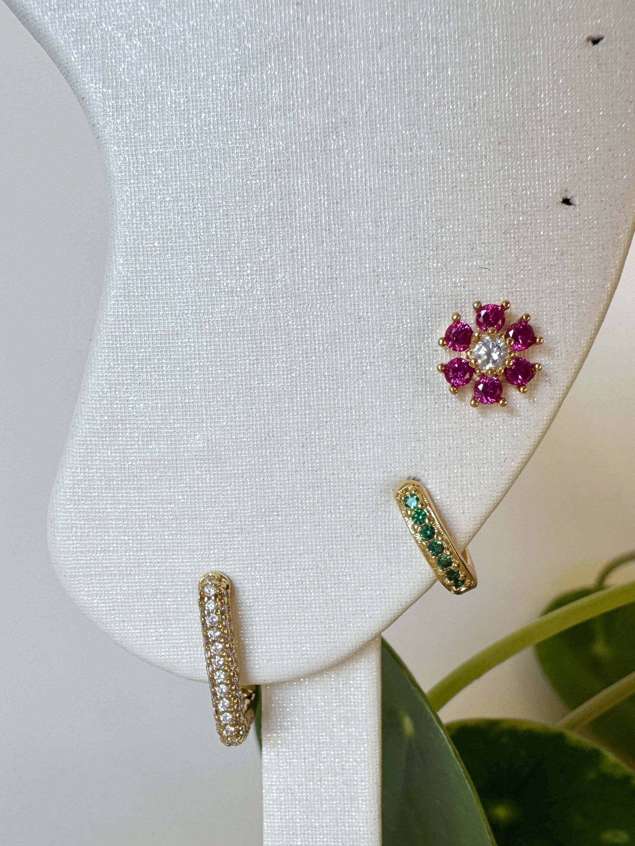 Gold earrings with gemstones on a white display stand with green leaves in the background