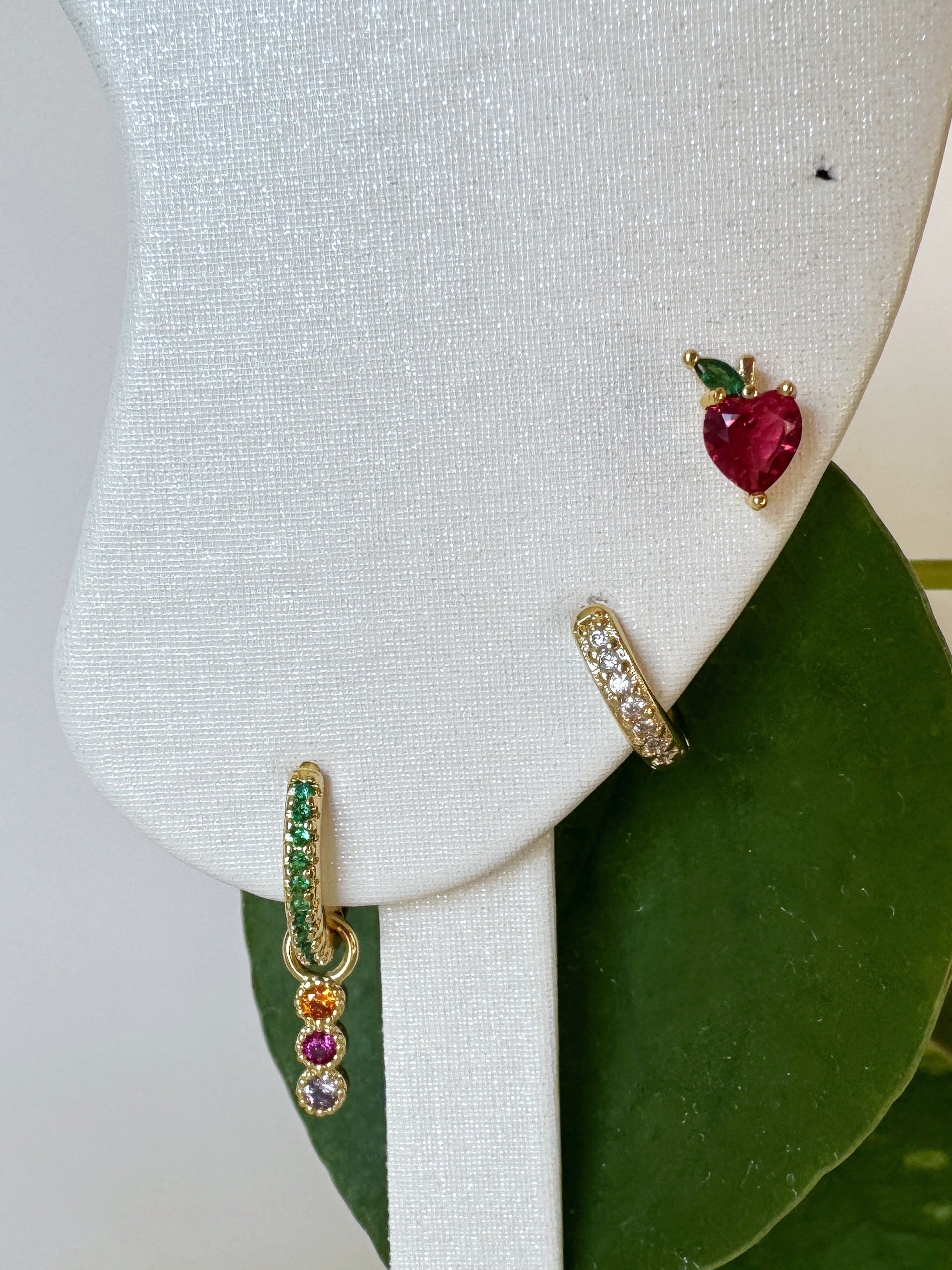 Pair of earrings in apple design with gemstones on a white stand with green leaves in the background
