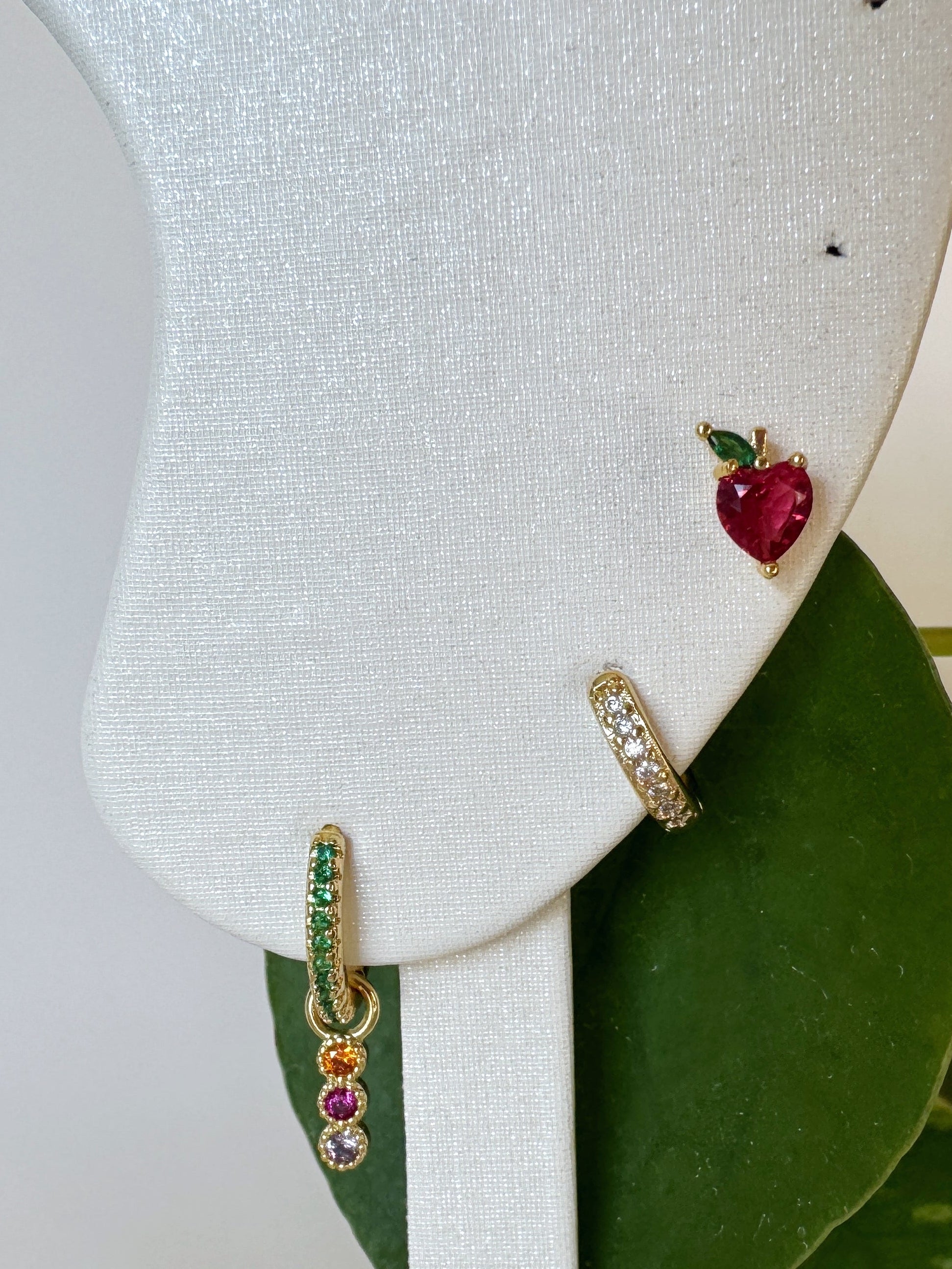 Pair of earrings with apple gemstones on a white stand with green leaves in the background. Classic gold hoops and multicolor