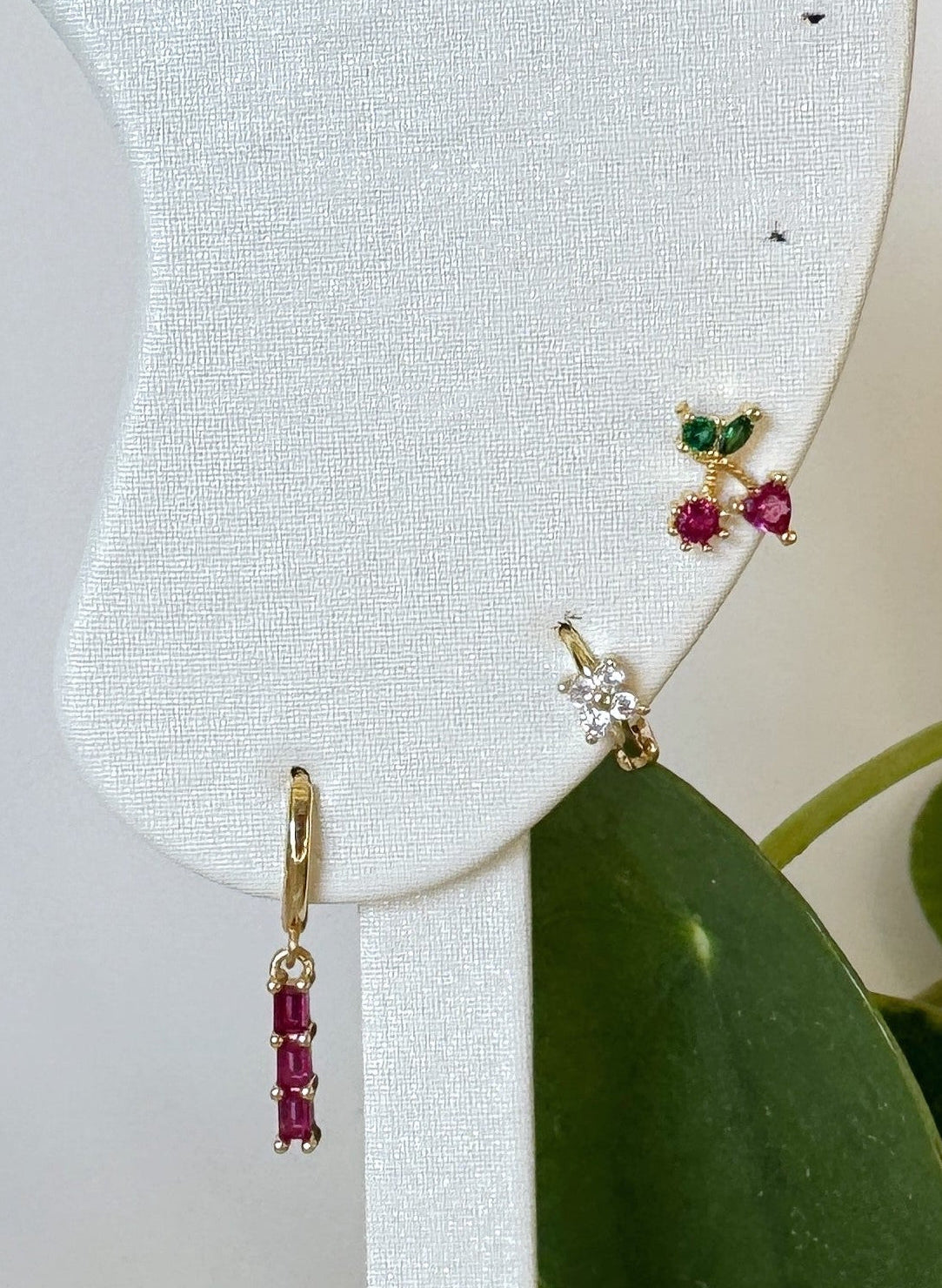 Gold earrings with red and green gemstones on a white earring display with a green leaf background.
