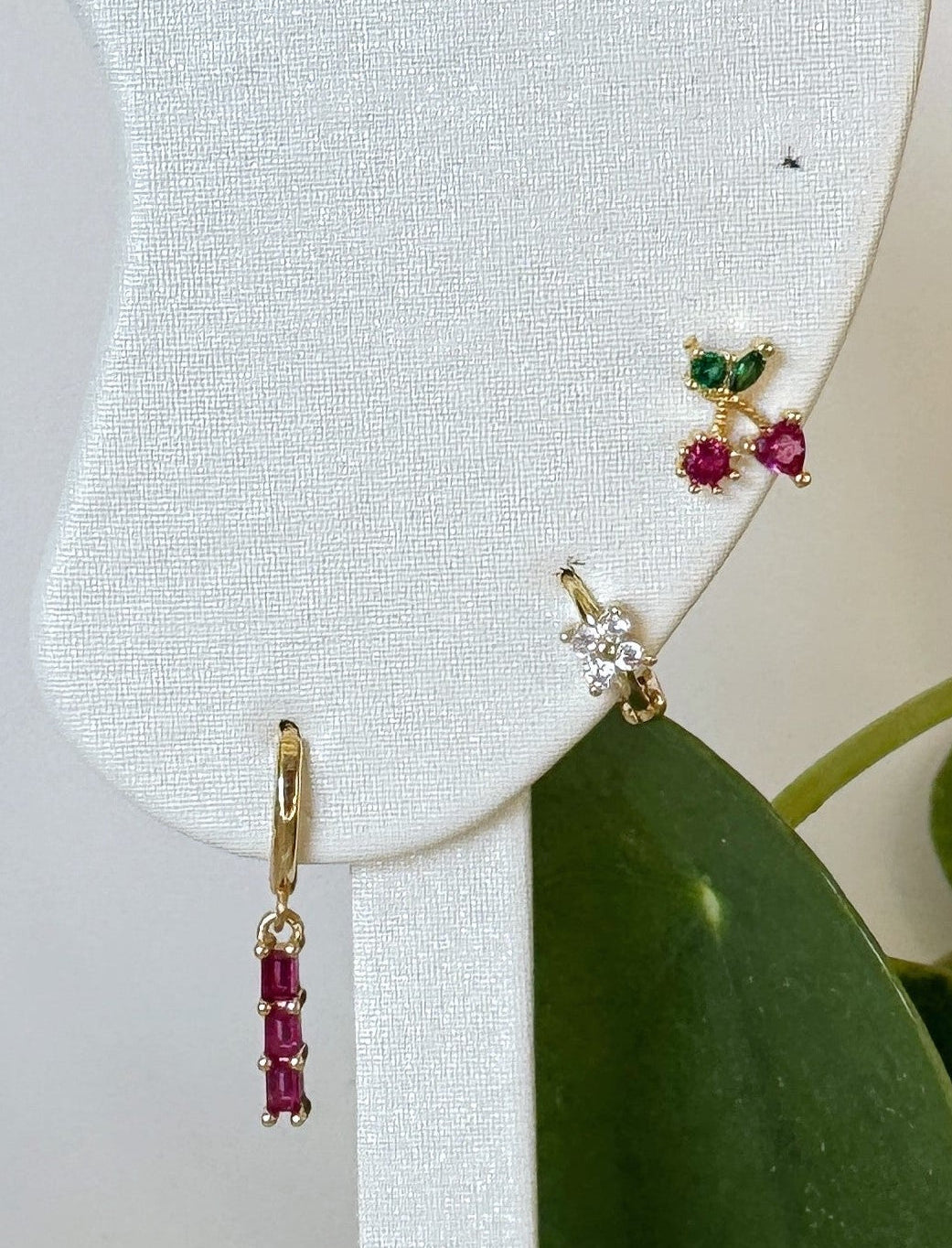 Gold earrings with red and green gemstones on a white earring display with a green leaf background.