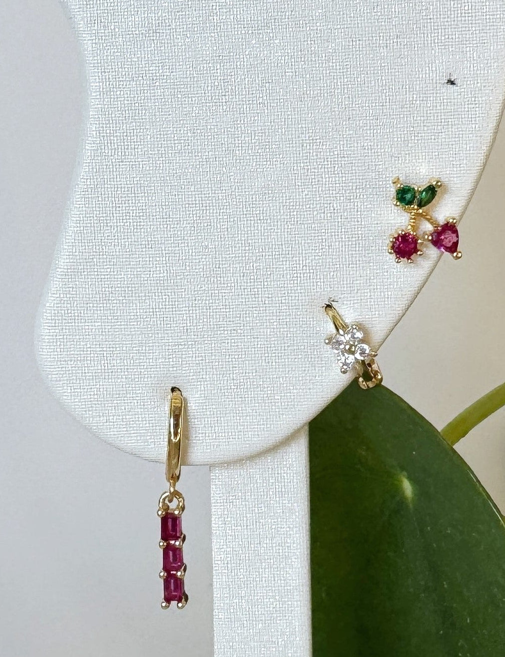 Gold earrings with red and green gemstones on a white earring display with a green leaf background.