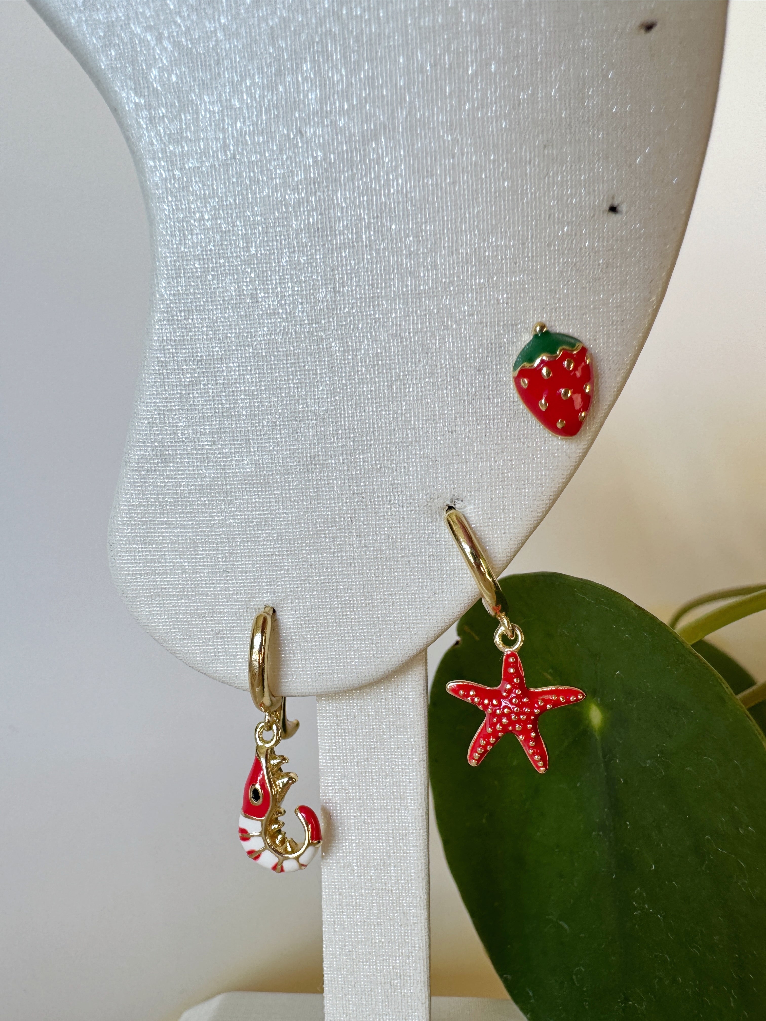 Three red earrings on a white stand with a green leaf background in shapes of strawberry, sea star and shrimp