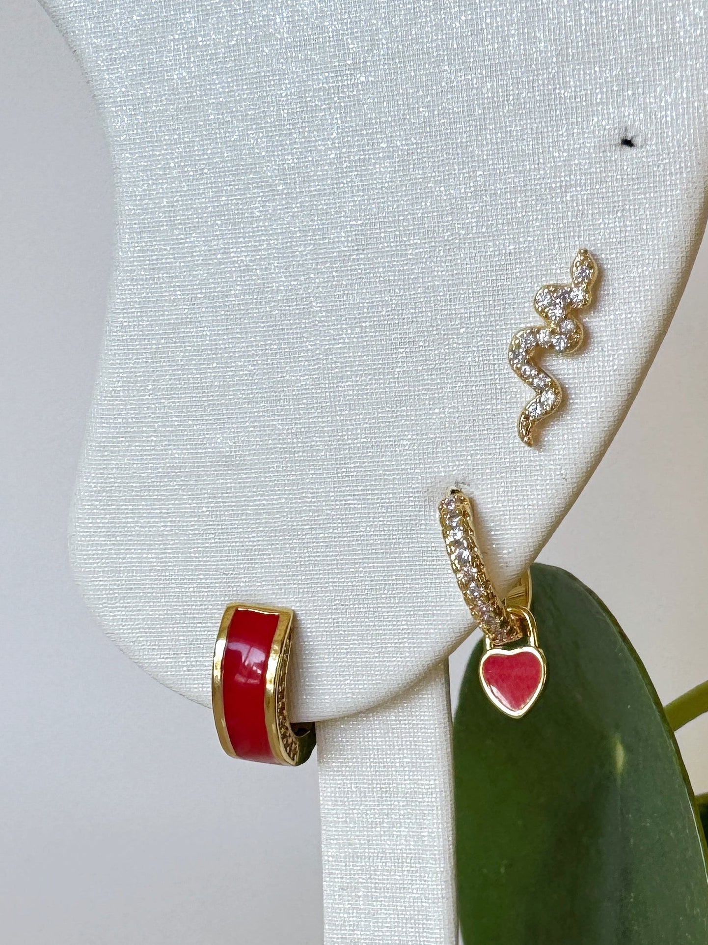 Earrings with snake and heart design in red accent colors on a white stand with green leaves in the foreground