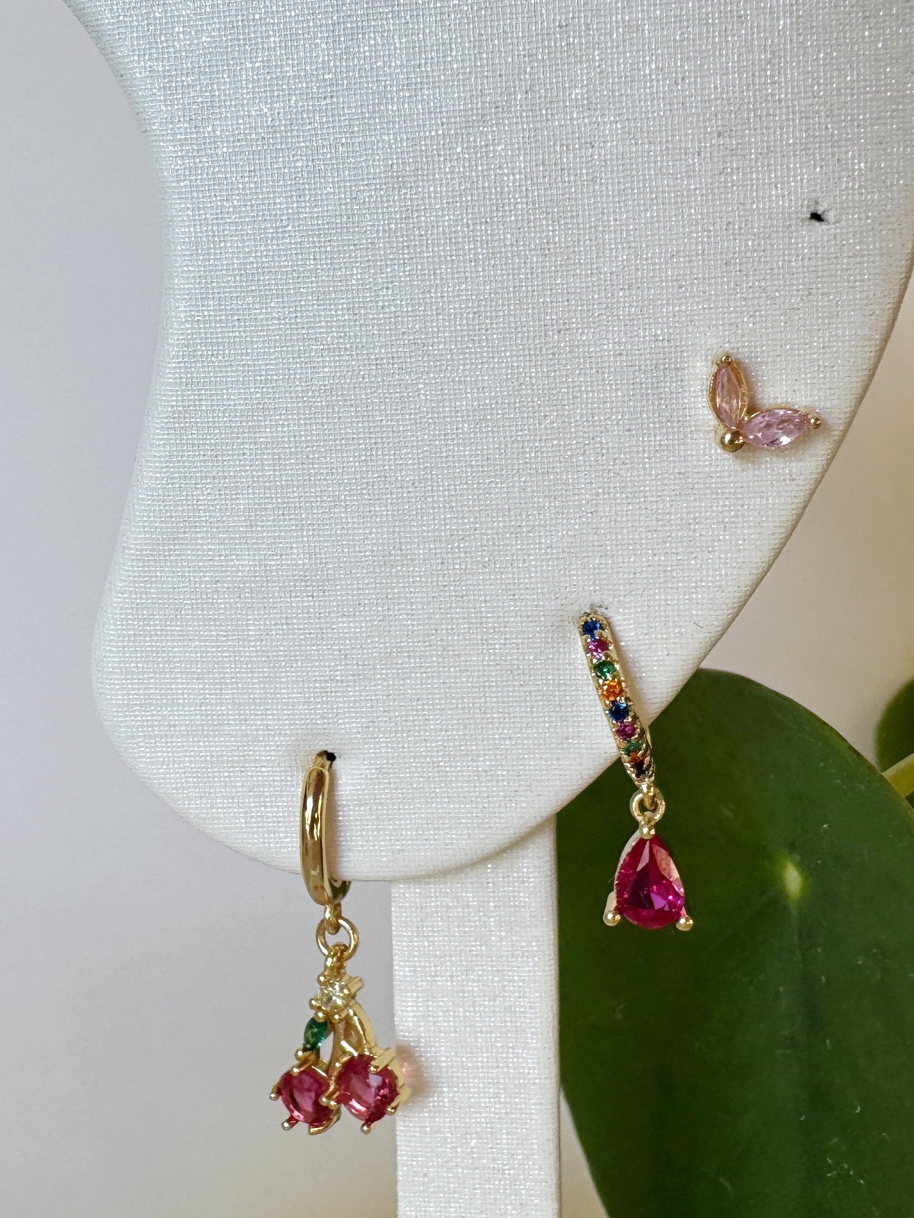 Set of gold earrings with colorful pink gemstones on a white jewelry display with green leaves in the background