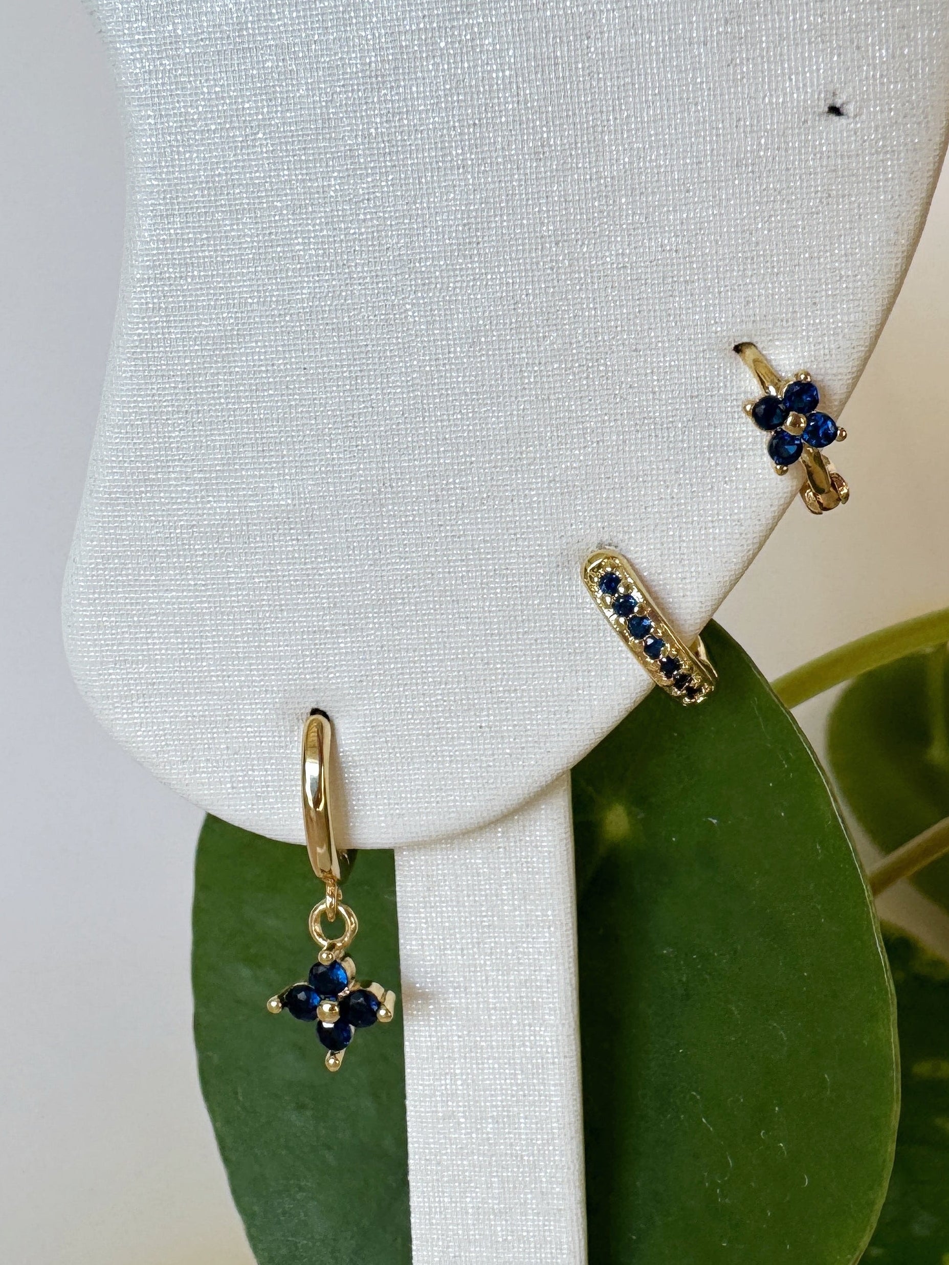Gold earrings with blue stones on a white stand with green leaves in the background