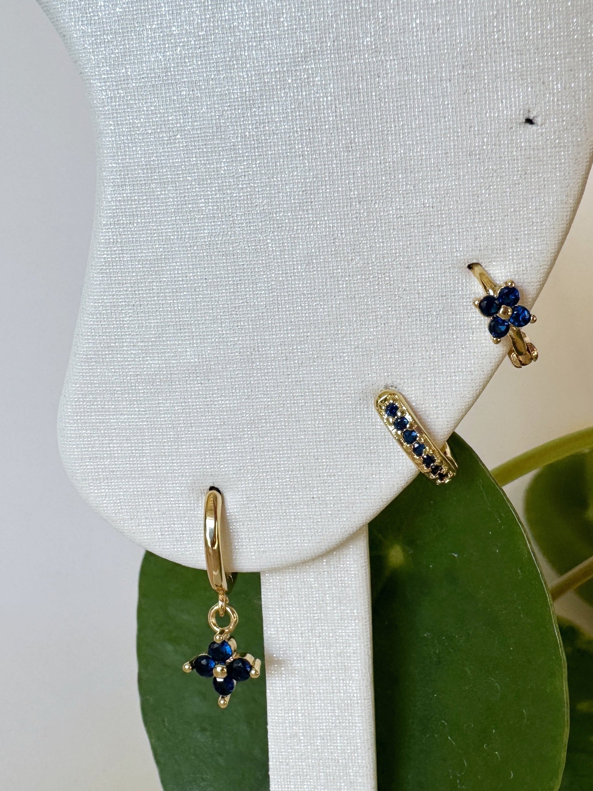 Gold earrings with sapphire blue stones on a white stand with green leaves in the background