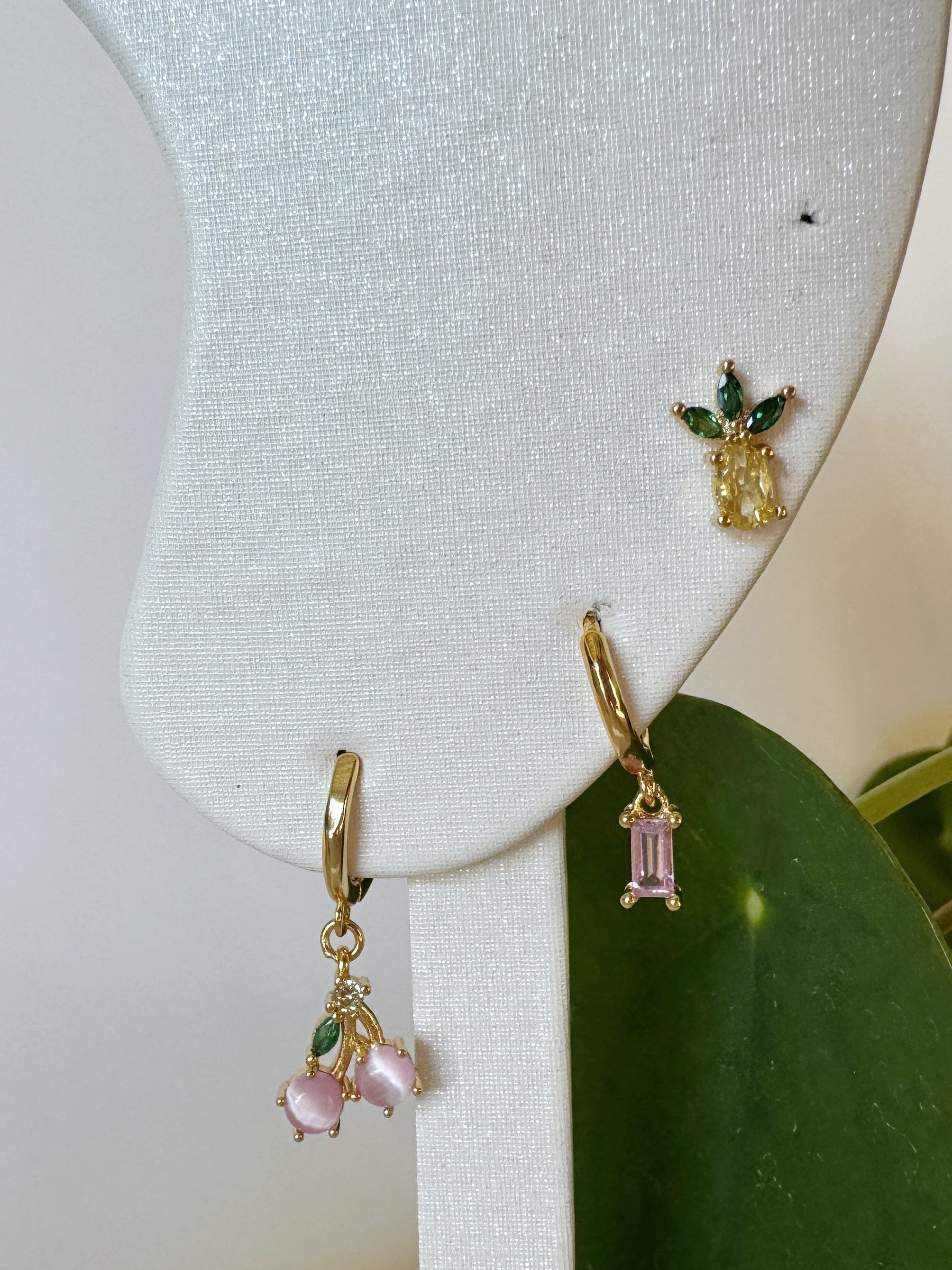 Gold earrings with pineapple and cherry design and with pink gemstones on a white stand with green leaves.