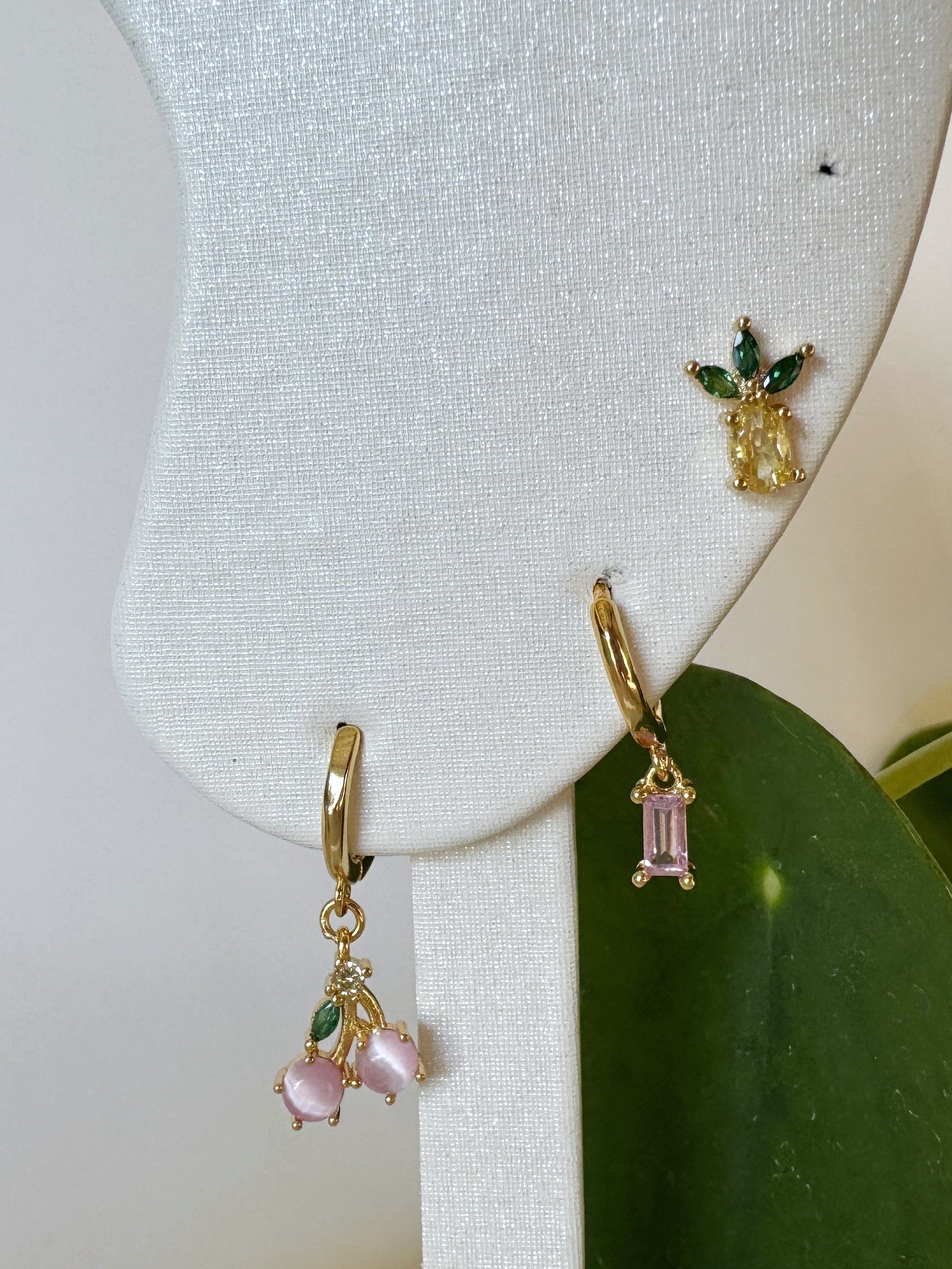 Gold earrings with pink gemstones on a white stand with green leaves. Pineapple and cherry design.