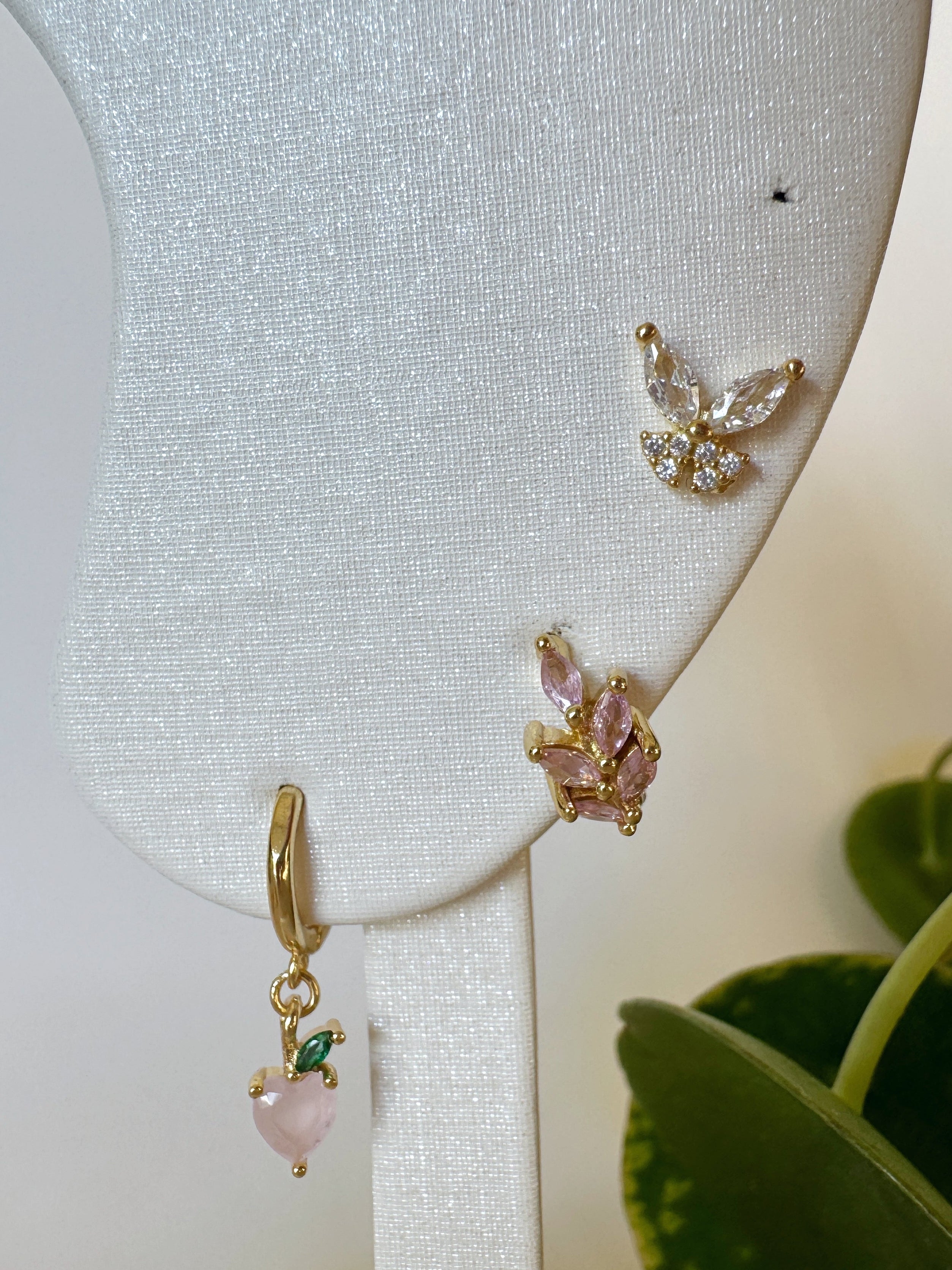 Gold butterfly and peach earrings with pink stones on a white stand with a plant in the background
